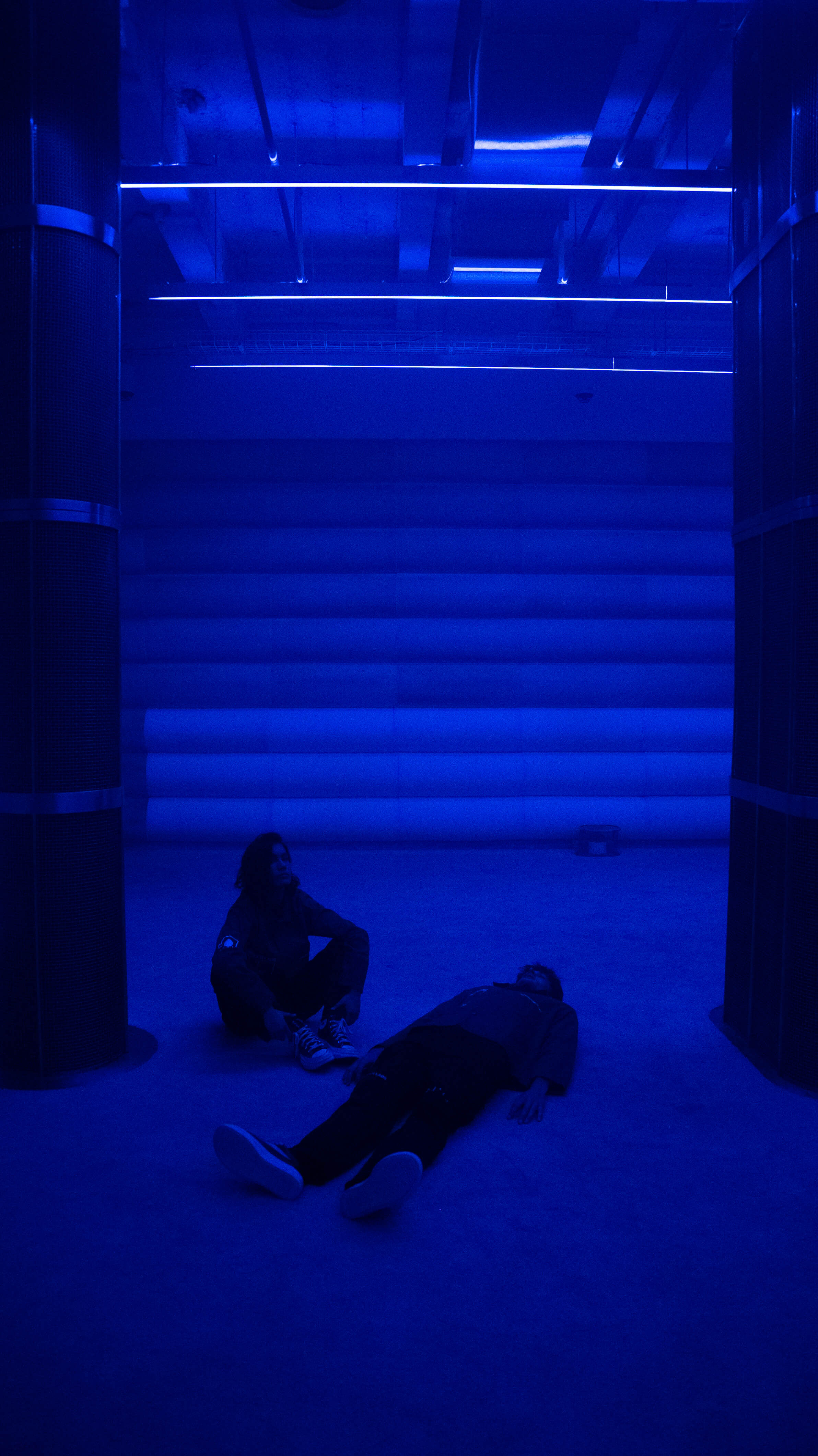 Two visitors rest on the floor of the 4DSound chamber at Mercer Labs, one lying down and one seated, under deep blue light and ceiling LED strips, Lower Manhattan.