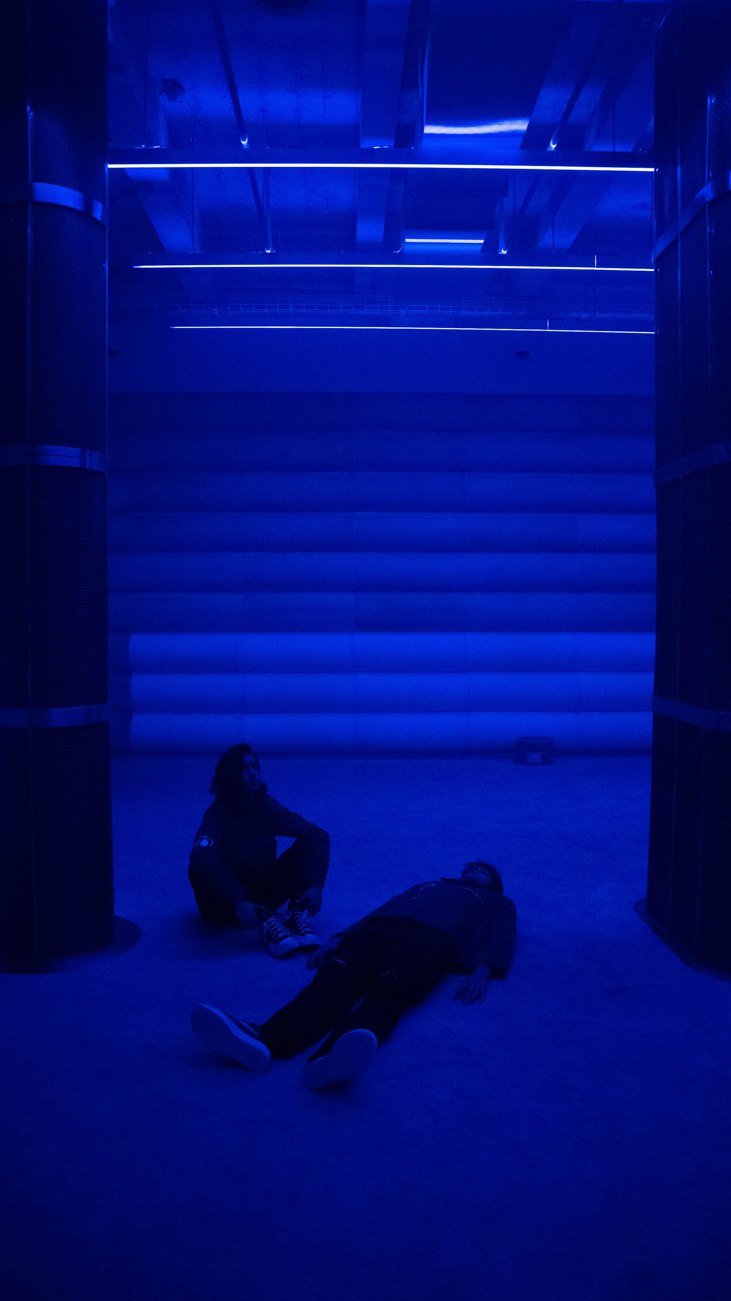 Two visitors rest on the floor of the 4DSound chamber at Mercer Labs, one lying down and one seated, under deep blue light and ceiling LED strips, Lower Manhattan.