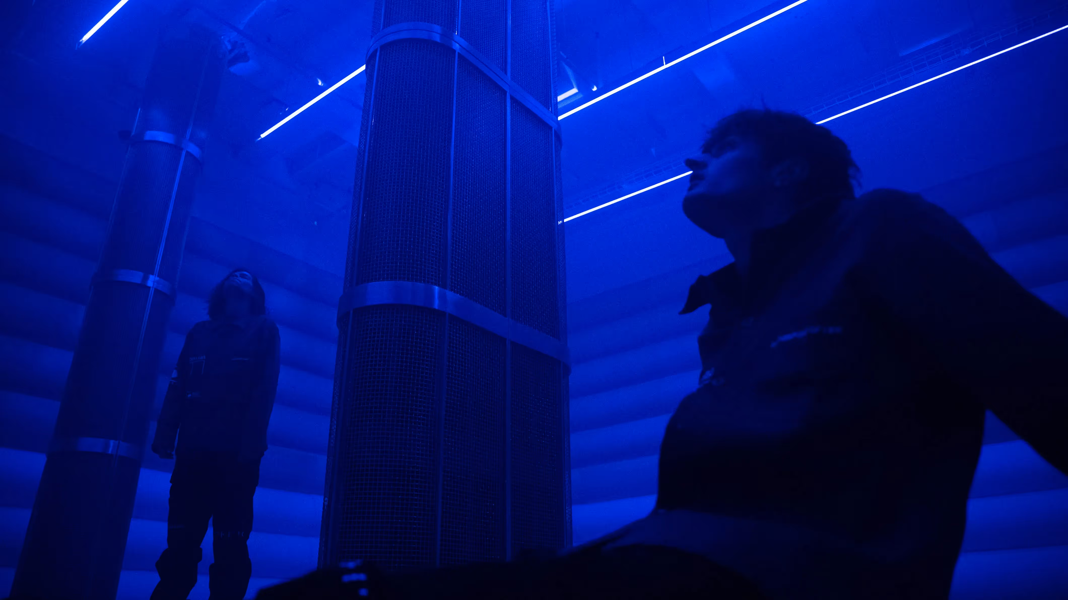 Low angle view of two visitors standing between tall mesh speaker pillars in the blue-lit 4DSound chamber at Mercer Labs, NYC.