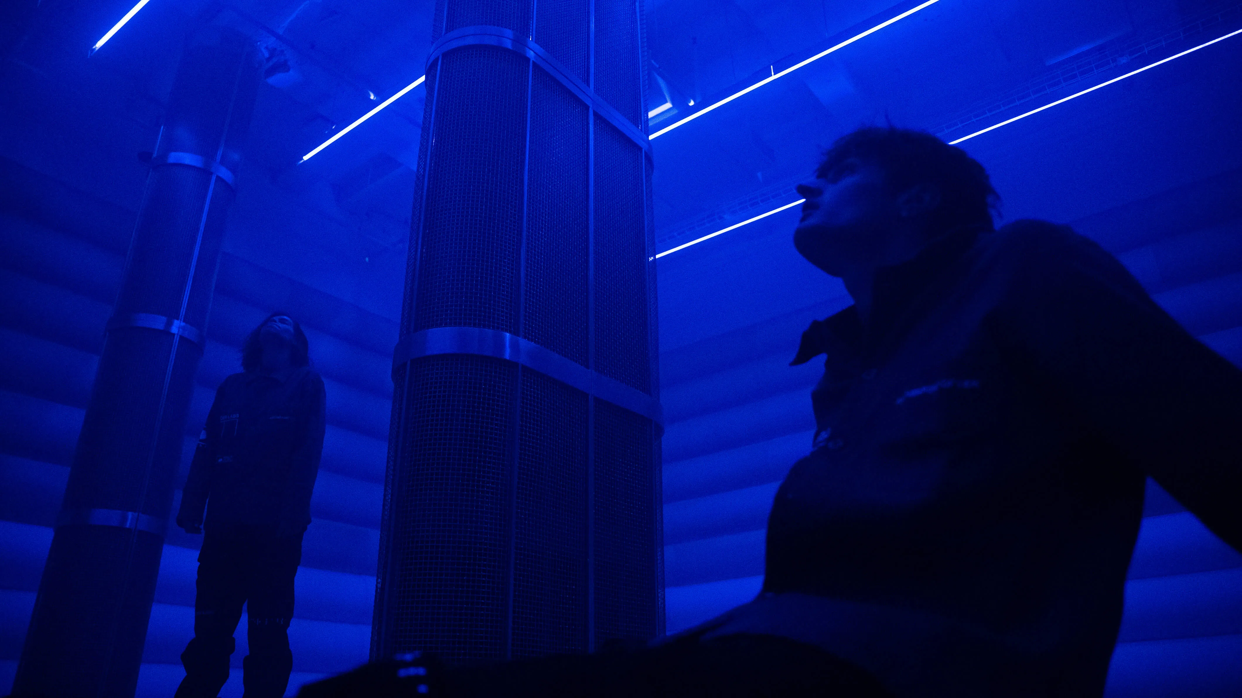 Low angle view of two visitors standing between tall mesh speaker pillars in the blue-lit 4DSound chamber at Mercer Labs, NYC.