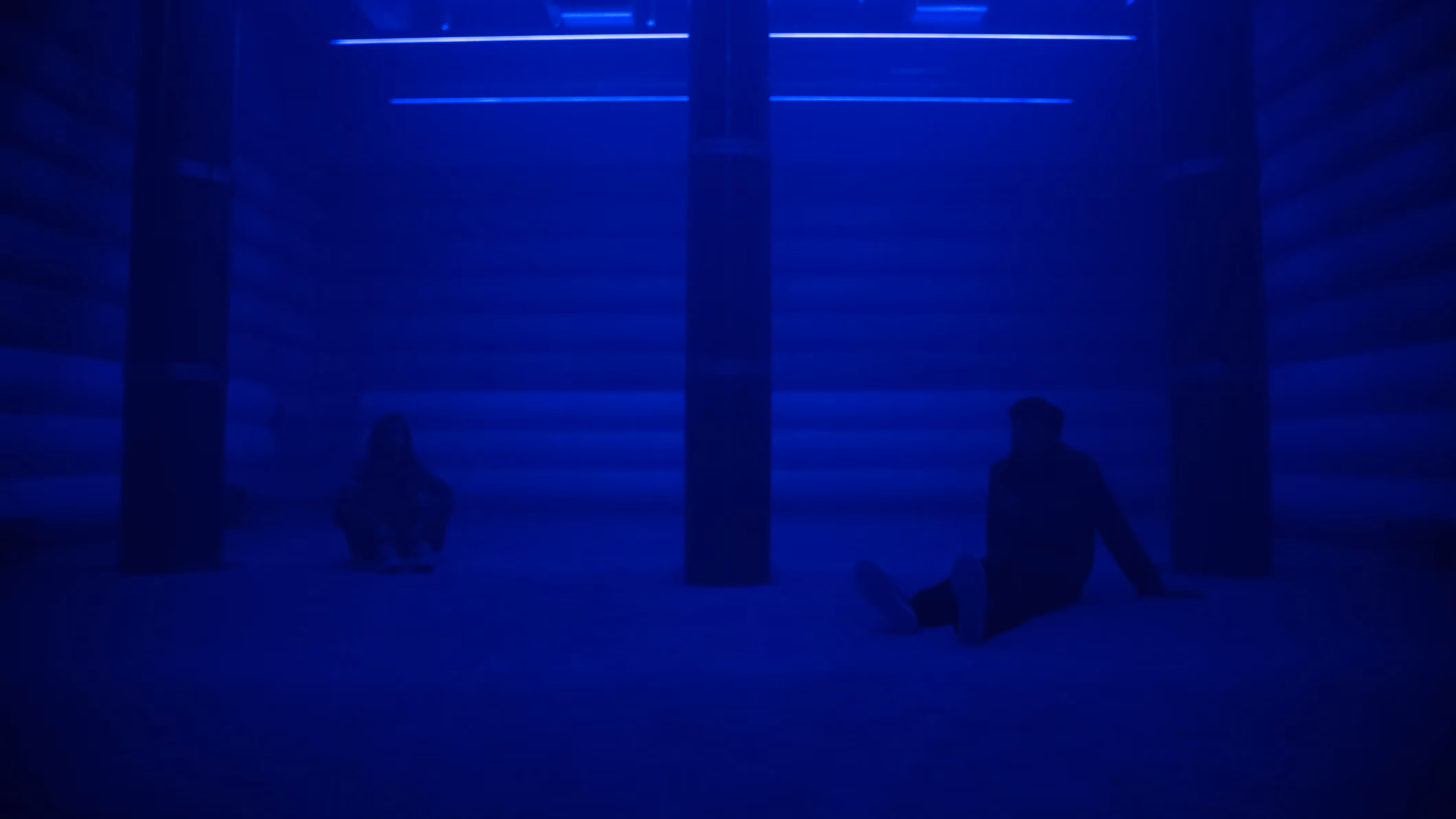 Two visitors sit on the floor of the 4DSound chamber at Mercer Labs, silhouetted in deep blue light between tall speaker columns, a museum in NYC.