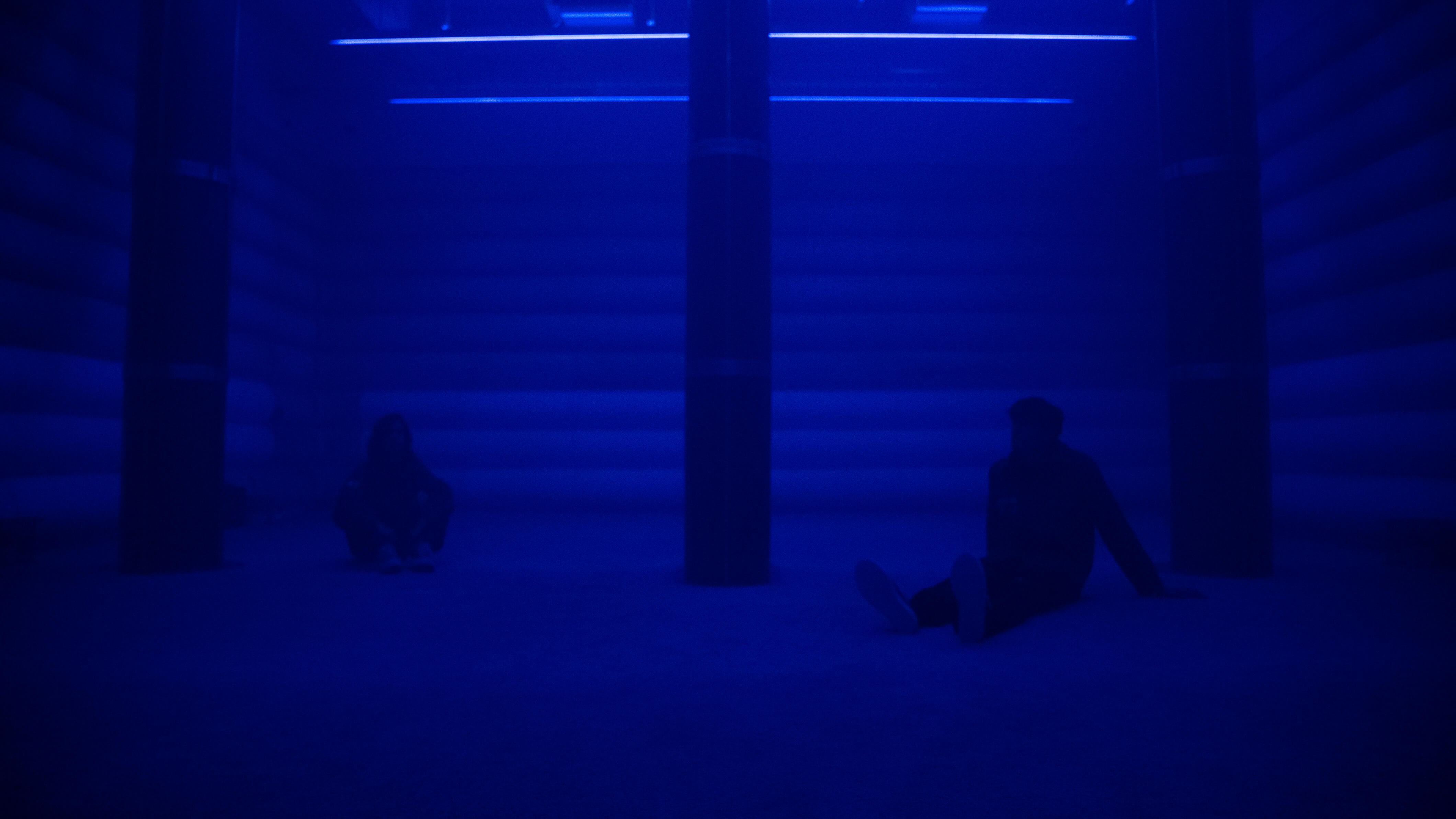 Two visitors sit on the floor of the 4DSound chamber at Mercer Labs, silhouetted in deep blue light between tall speaker columns, a museum in NYC.