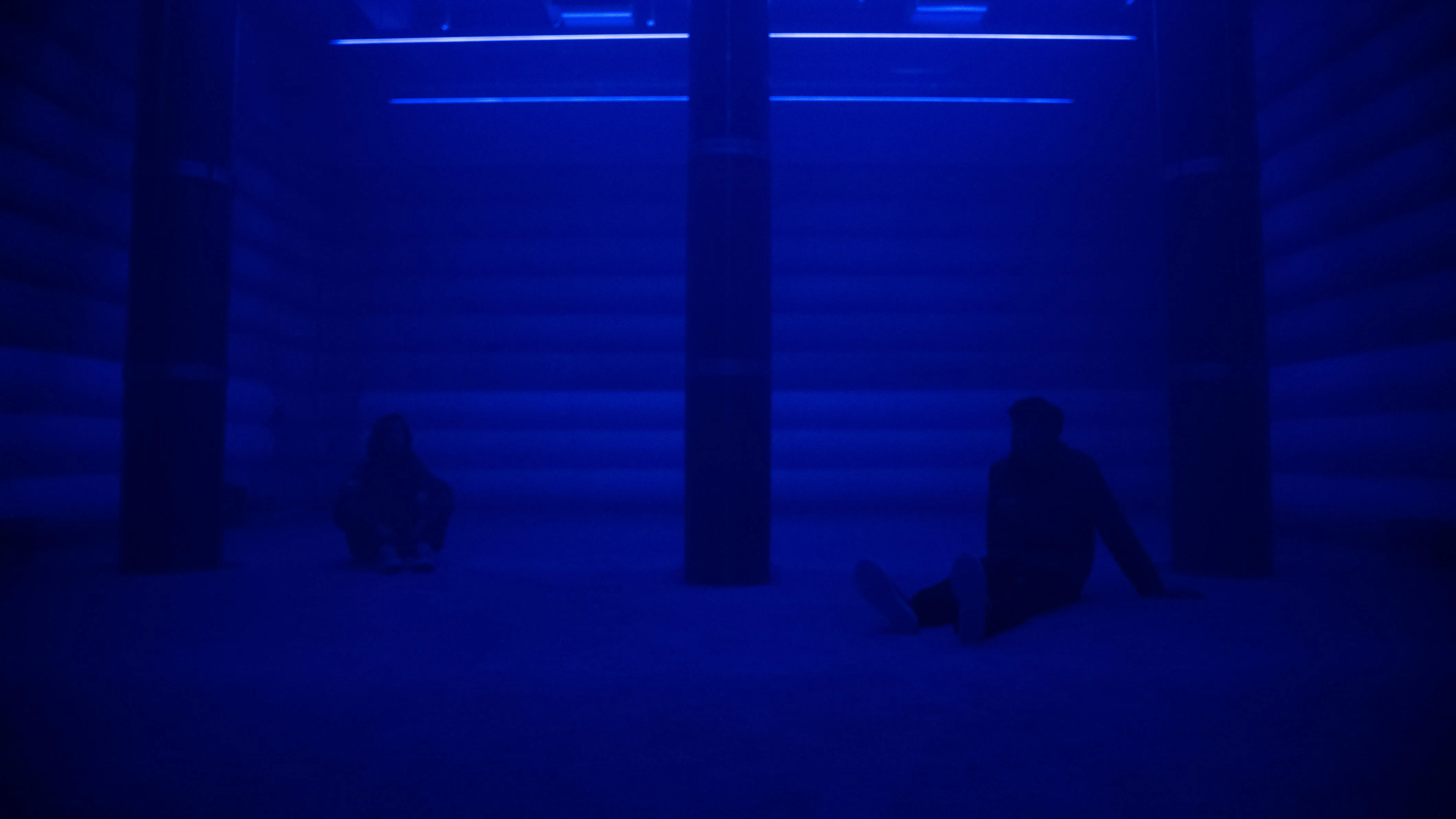 Two visitors sit on the floor of the 4DSound chamber at Mercer Labs, silhouetted in deep blue light between tall speaker columns, a museum in NYC.