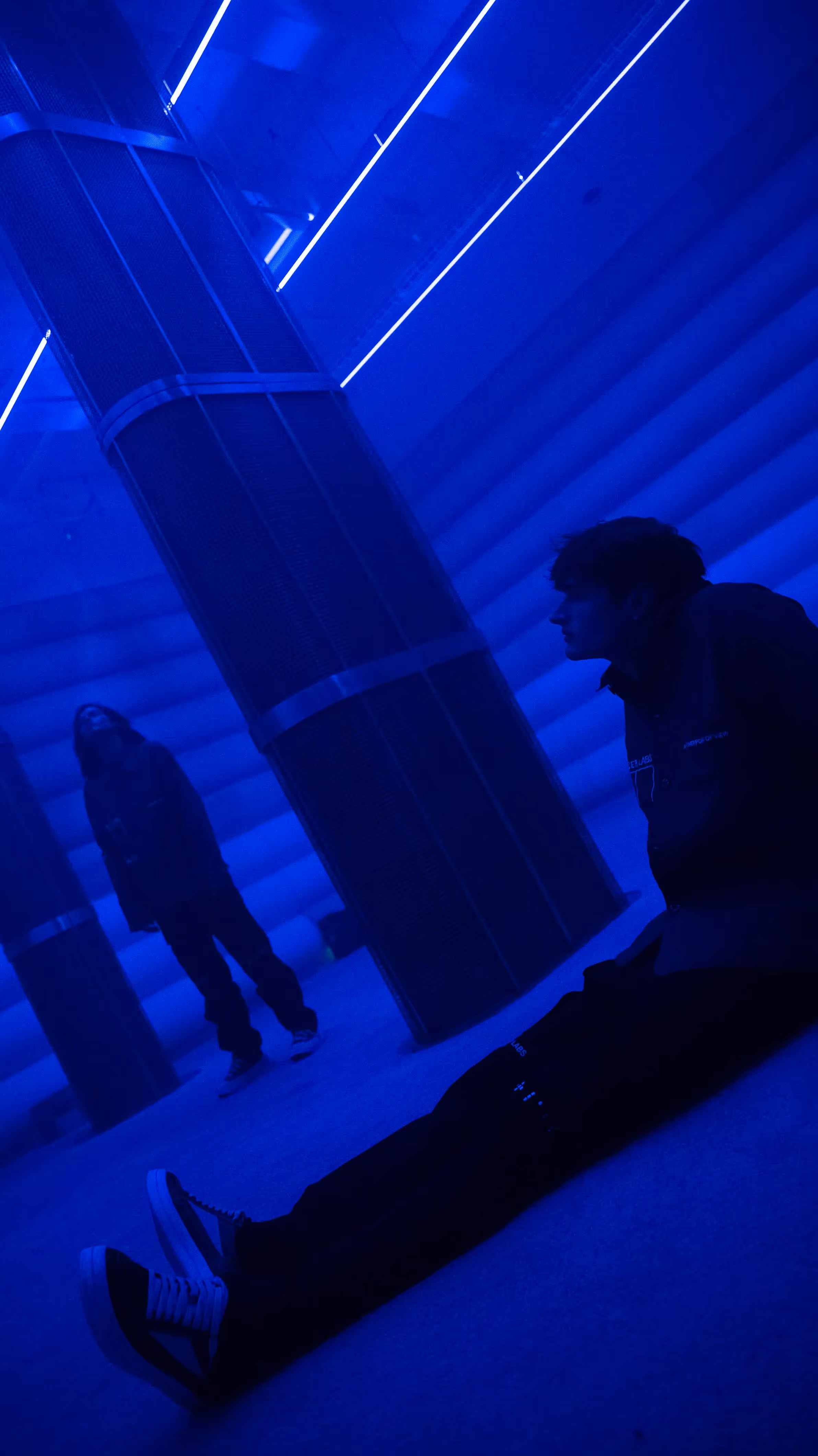Visitor in a Mercer Labs jacket sits on the floor beside a mesh speaker column with their reflection visible in the blue 4DSound chamber at Mercer Labs, a museum in New York.