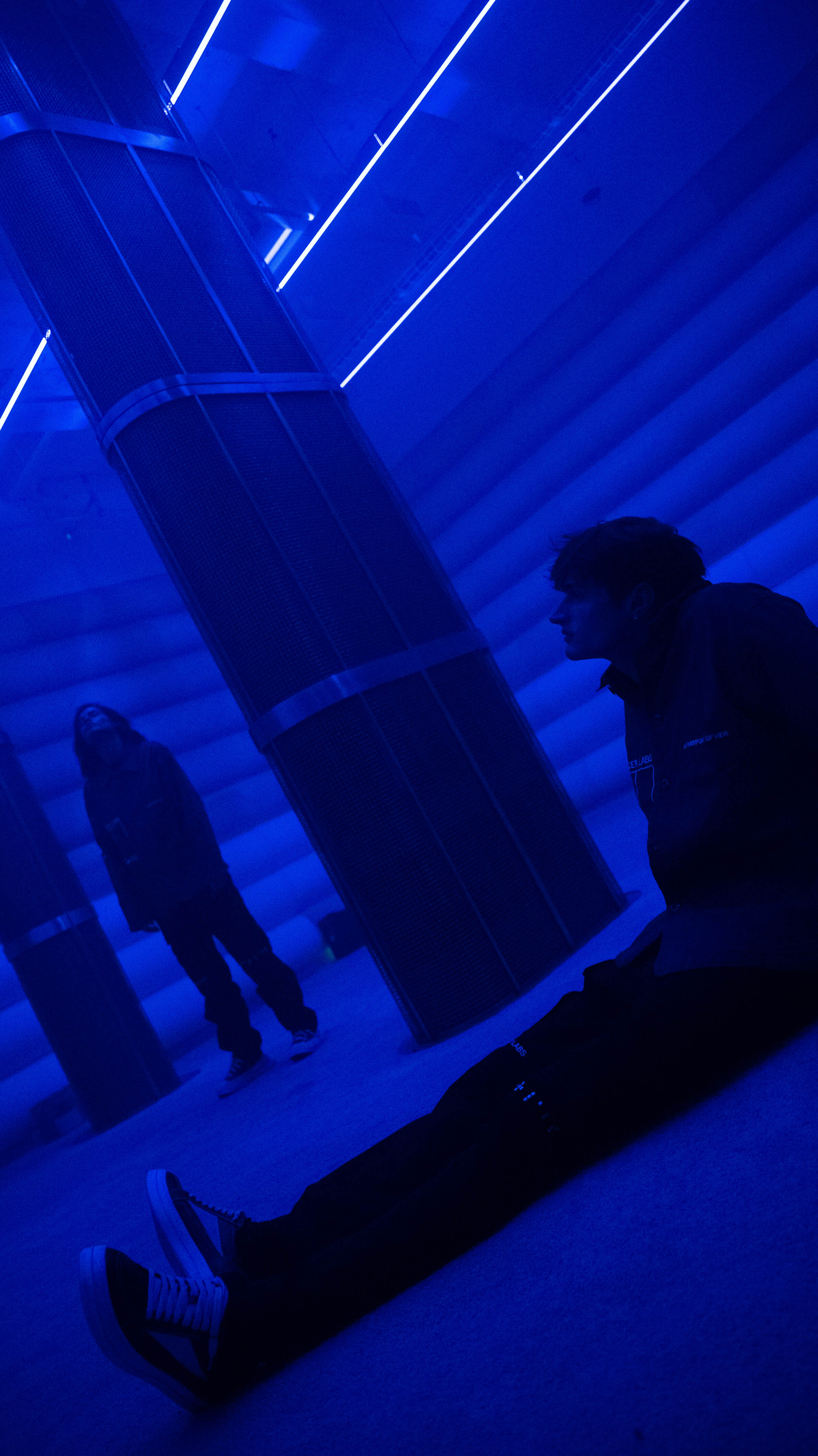 Visitor in a Mercer Labs jacket sits on the floor beside a mesh speaker column with their reflection visible in the blue 4DSound chamber at Mercer Labs, a museum in New York.