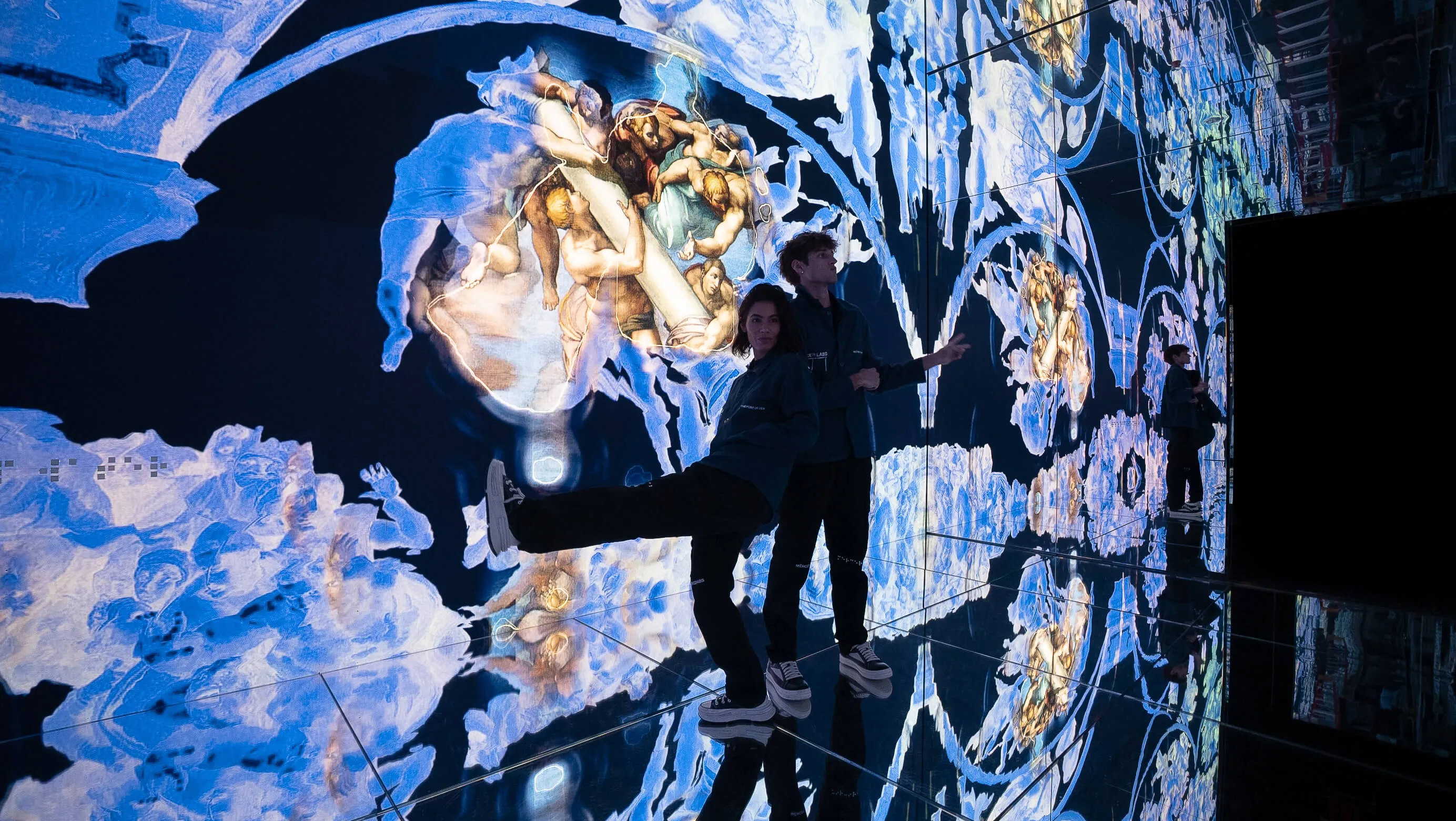 Two visitors pose playfully before cascading blue projections of Michelangelo figures reflected across the mirrored floor and walls of Infinite at Mercer Labs, Lower Manhattan.
