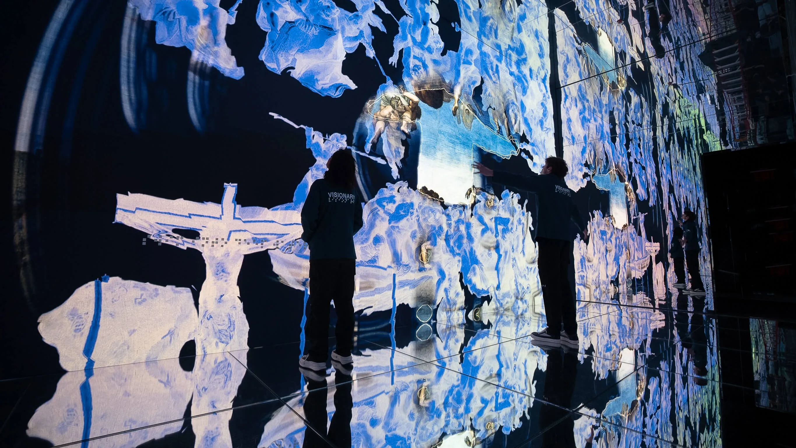 Two visitors in Visionary jackets stand inside the mirrored Infinite gallery at Mercer Labs, surrounded by towering fragmented projections of classical figures, NYC.