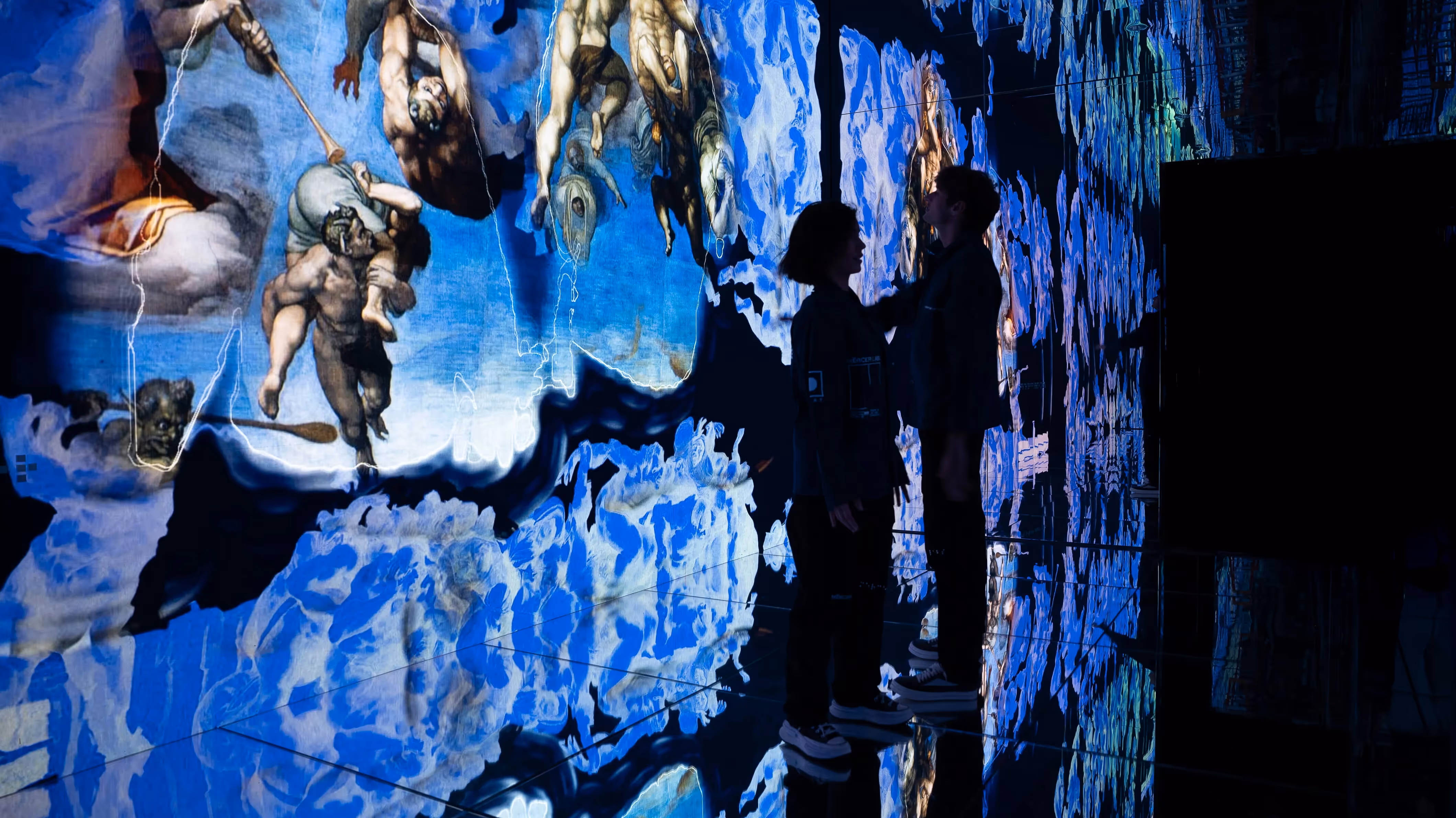Two visitors stand close together before a mirrored wall of shifting Michelangelo figures dissolving into blue fragments at Infinite, Mercer Labs, a museum in NYC.