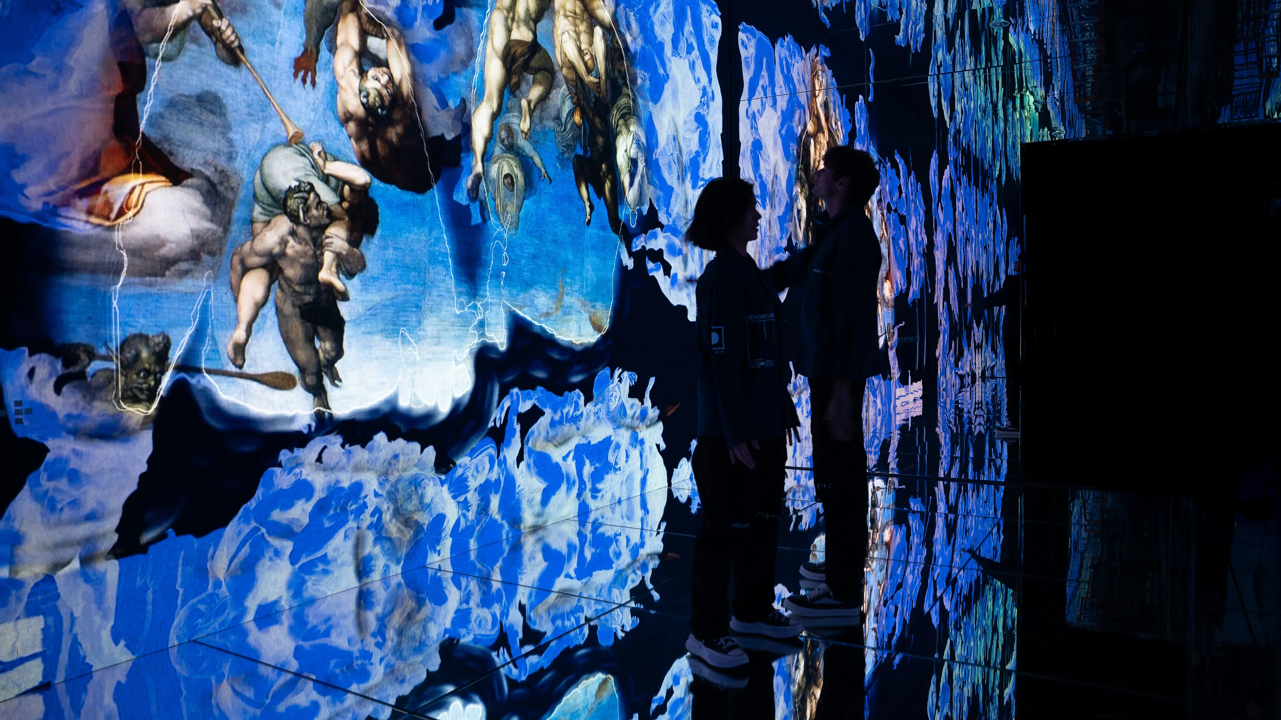 Two visitors stand close together before a mirrored wall of shifting Michelangelo figures dissolving into blue fragments at Infinite, Mercer Labs, a museum in NYC.
