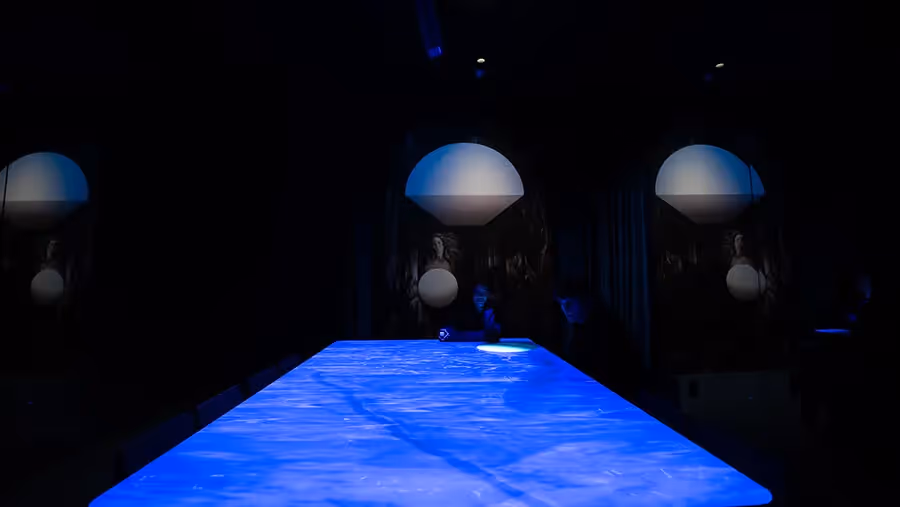 Visitors silhouetted at the glowing blue water-textured table of the Blue Room, with Venus paintings and dome forms repeated across the mirrors at Mercer Labs, an art and science museum.