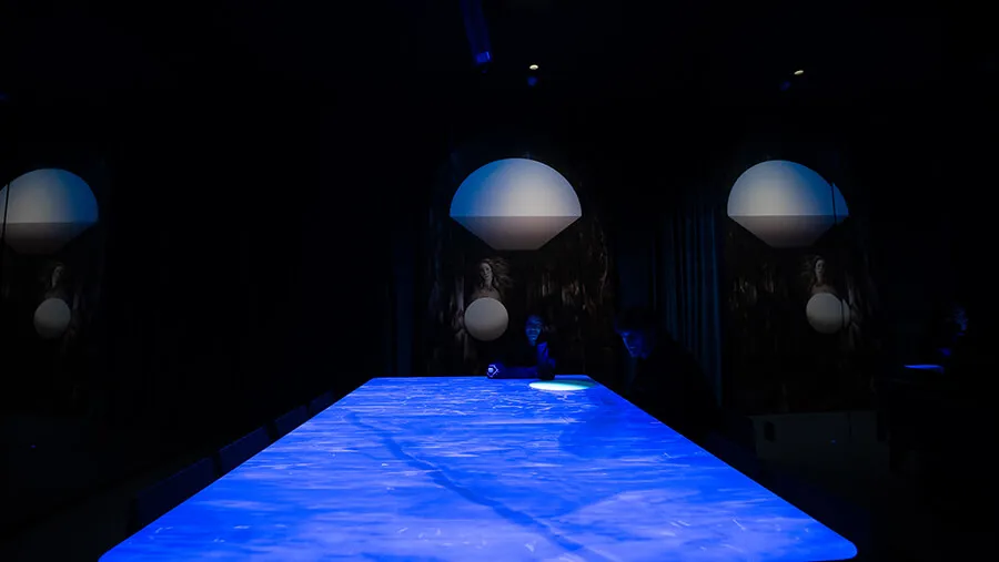 Visitors silhouetted at the glowing blue water-textured table of the Blue Room, with Venus paintings and dome forms repeated across the mirrors at Mercer Labs, an art and science museum.