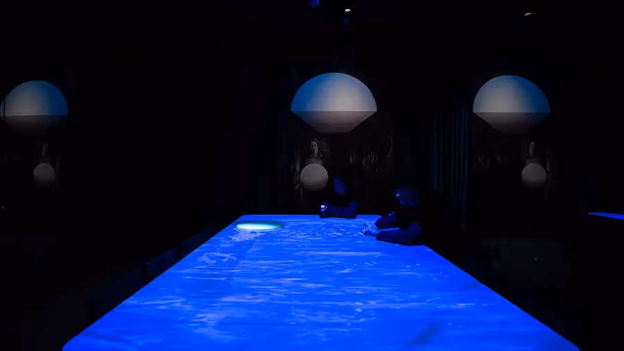 Wide view down a glowing blue table at the Blue Room with two visitors writing, flanked by Venus paintings and dome lights at Mercer Labs, a museum in NYC.