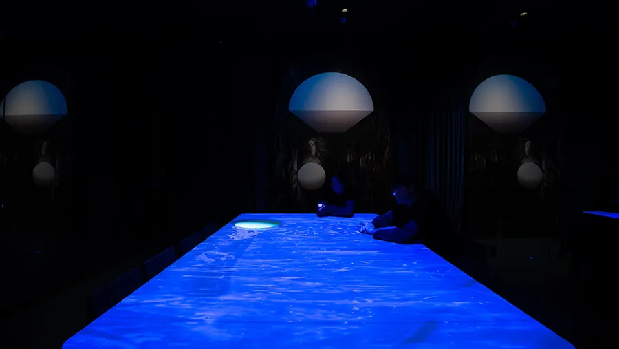 Wide view down a glowing blue table at the Blue Room with two visitors writing, flanked by Venus paintings and dome lights at Mercer Labs, a museum in NYC.