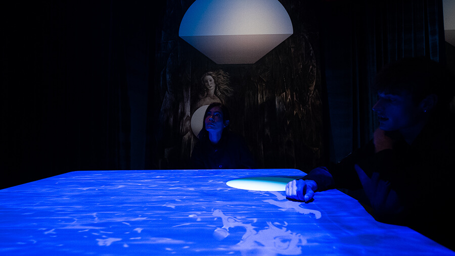Two visitors at the Blue Room table with a glowing orb of light on the surface and a classical Venus painting behind them at Mercer Labs, a museum of art and science in New York.