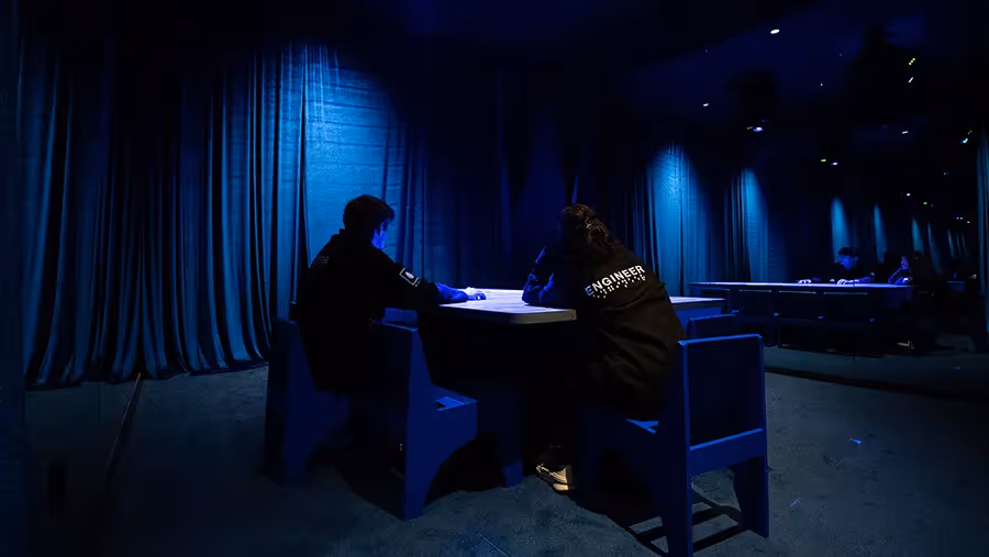 Two visitors in Mercer Labs Engineer jackets writing at a Blue Room table surrounded by dark blue velvet curtains at Mercer Labs, an immersive museum in Lower Manhattan.