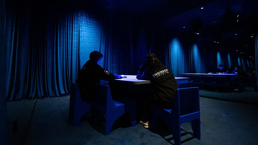 Two visitors in Mercer Labs Engineer jackets writing at a Blue Room table surrounded by dark blue velvet curtains at Mercer Labs, an immersive museum in Lower Manhattan.