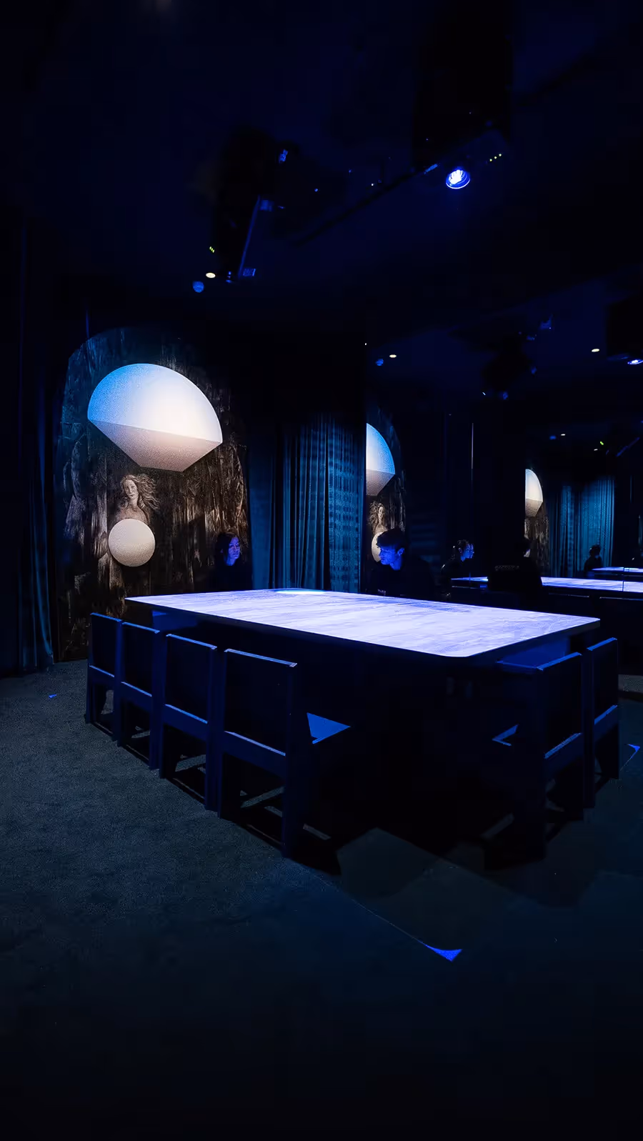 Tall view of an empty glowing blue table with overhead spotlights and a classical Venus painting on the wall at the Blue Room, Mercer Labs NYC.