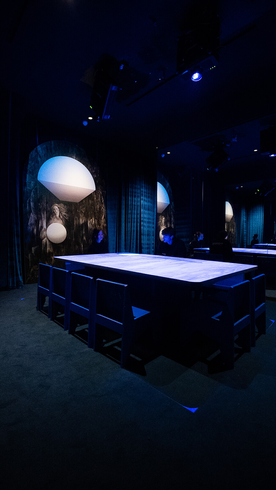Tall view of an empty glowing blue table with overhead spotlights and a classical Venus painting on the wall at the Blue Room, Mercer Labs NYC.