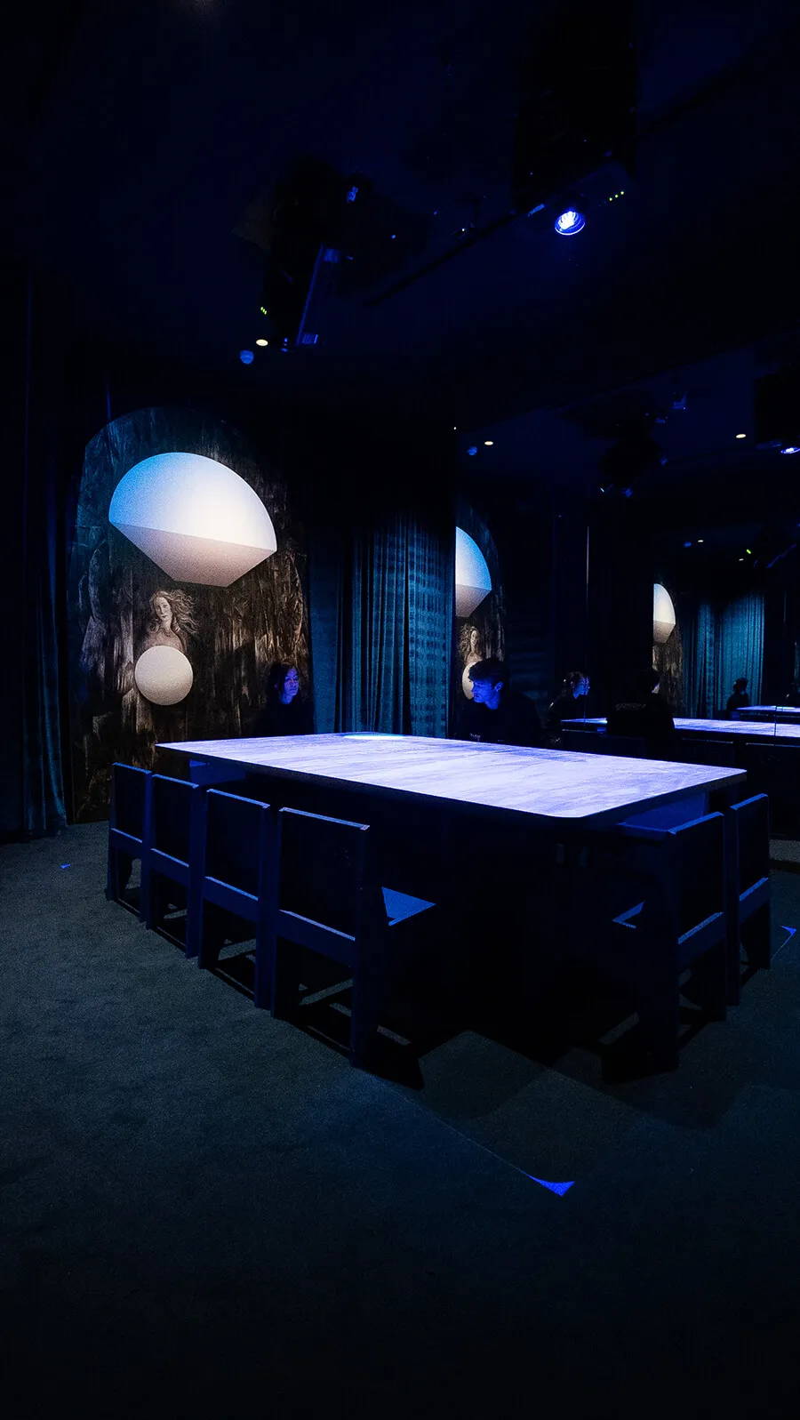 Tall view of an empty glowing blue table with overhead spotlights and a classical Venus painting on the wall at the Blue Room, Mercer Labs NYC.