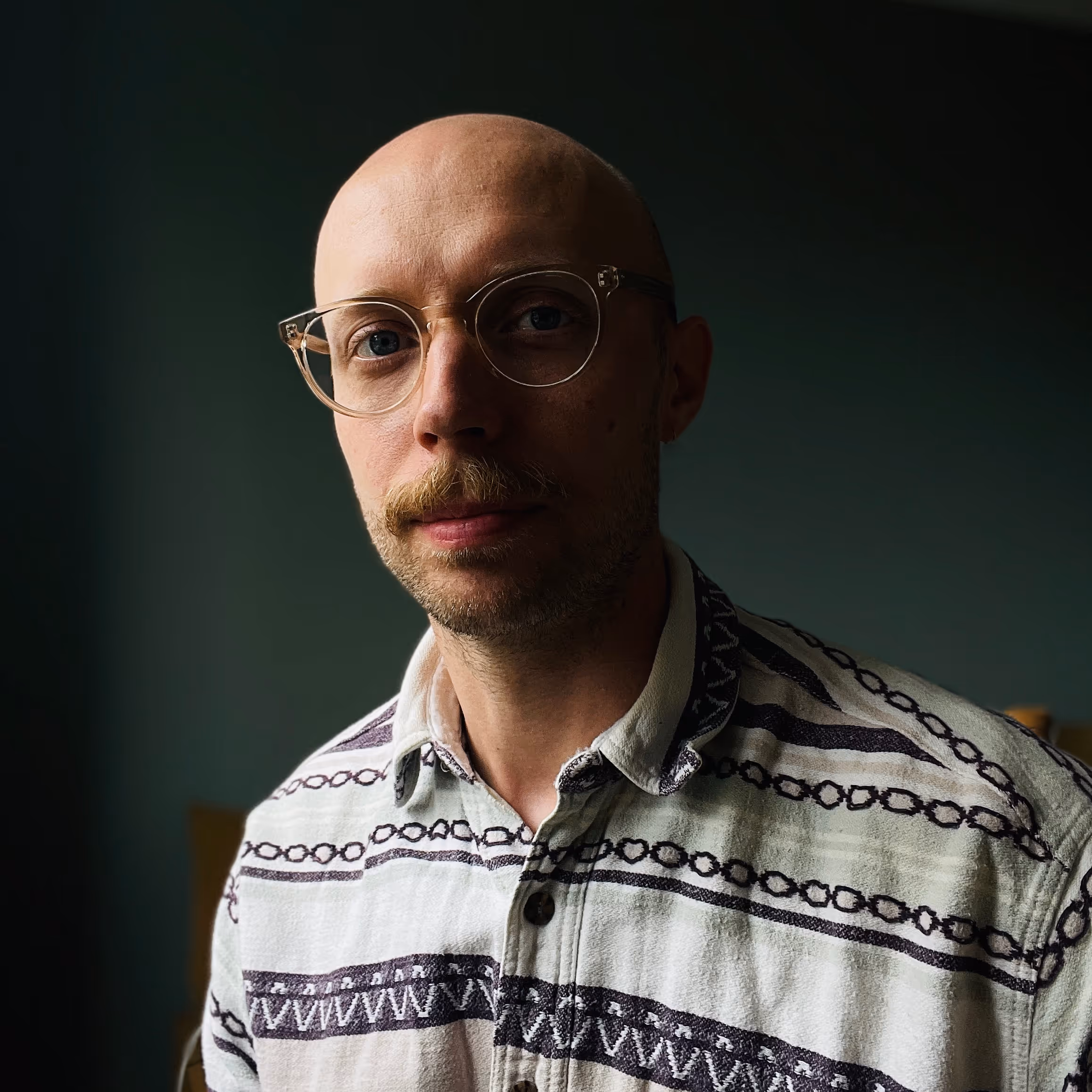 Portrait of a bald man with glasses and a mustache wearing a patterned button-up shirt.