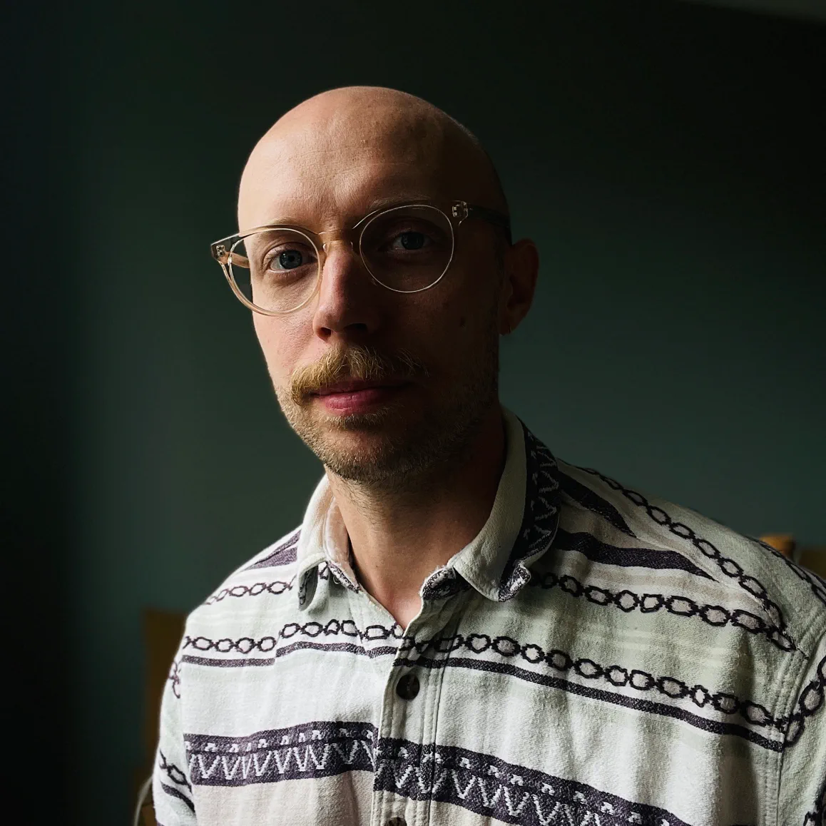 Portrait of a bald man with glasses and a mustache wearing a patterned button-up shirt.