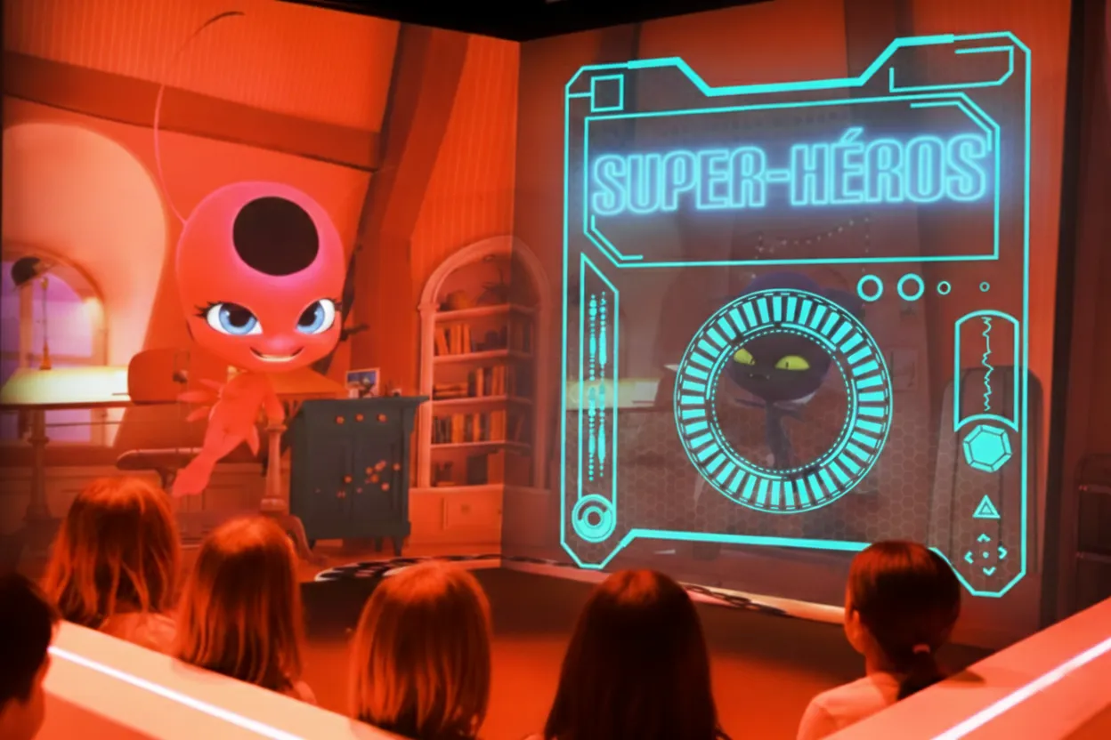 Children watching a holographic animation of a pink superhero character and a digital interface labeled 'SUPER-HÉROS' in a room with orange lighting.