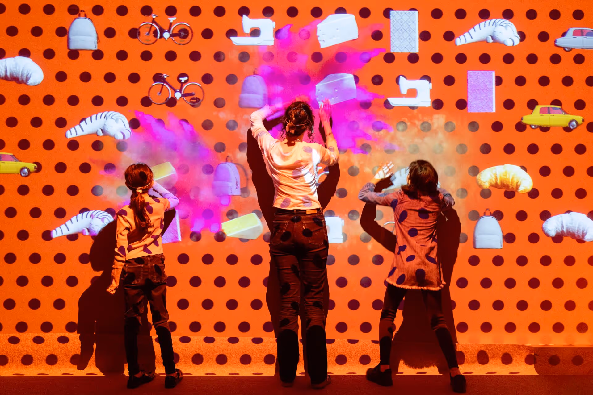 Children and adults playing a ball toss game in a bright red room with a large screen displaying the text 'Toucher les cibles avec les balles'.