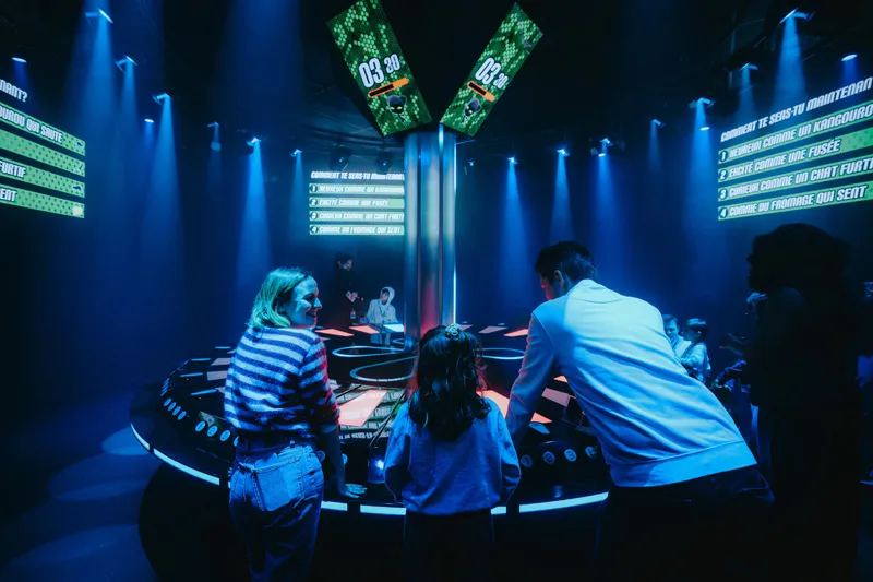 Children and adults standing and running on a pink-lit stage with hexagonal wall panels, with large text 'RÉFLEXION RAPIDE' and a green stopwatch icon in the center.