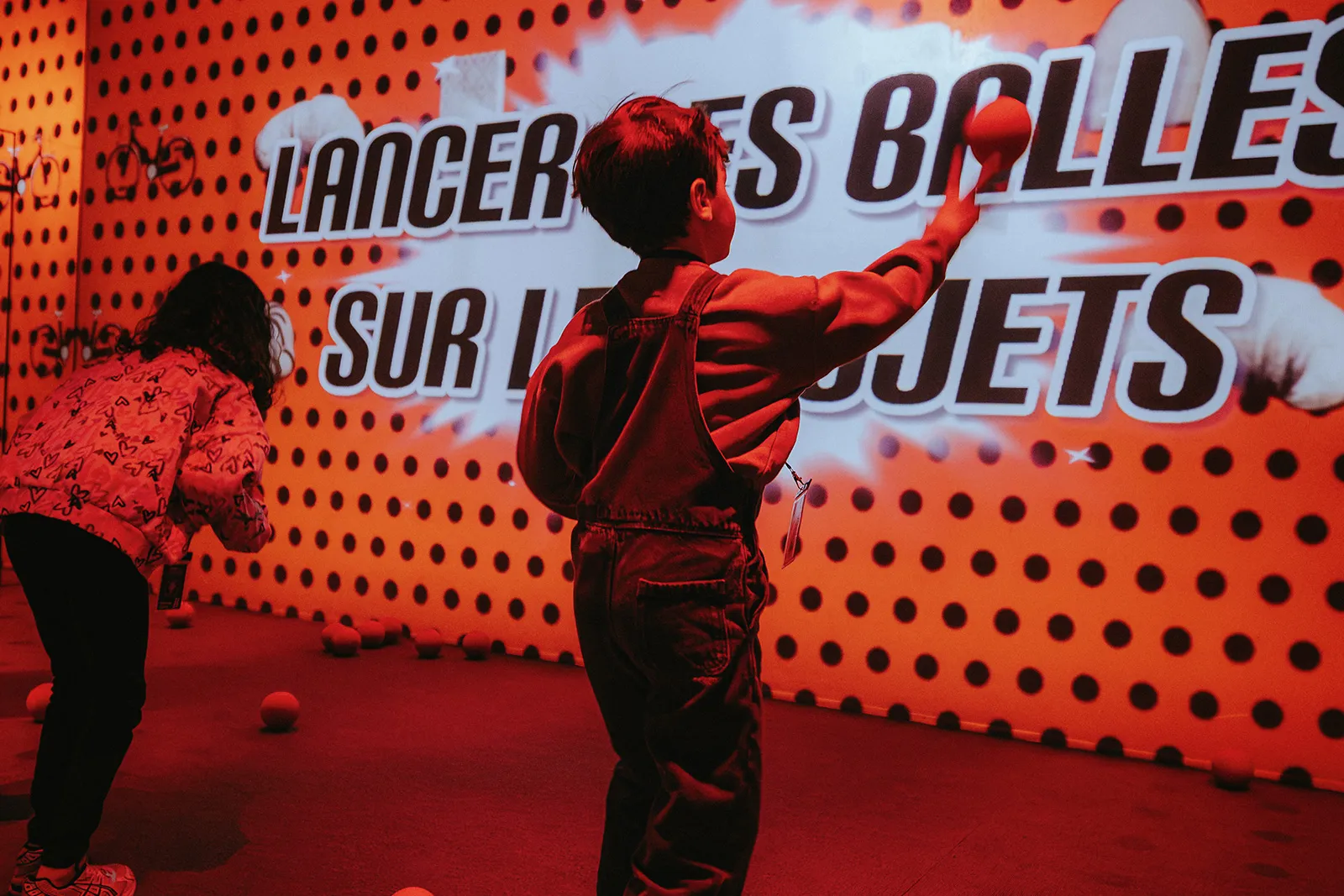 Children and adults playing a ball toss game in a bright red room with a large screen displaying the text 'Toucher les cibles avec les balles'.