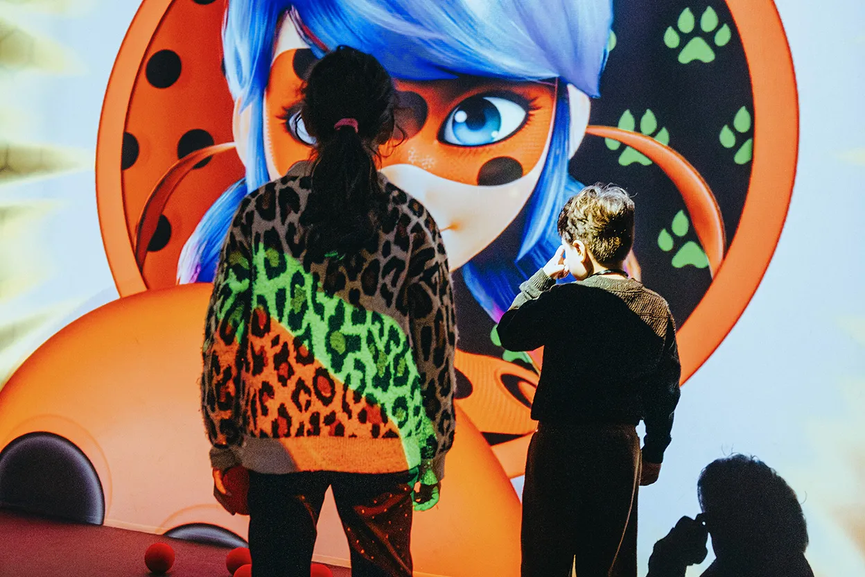 Audience watching a show with animated characters Chat Noir and Ladybug against green and red spotlights and the text 'Paris a besoin de toi'.