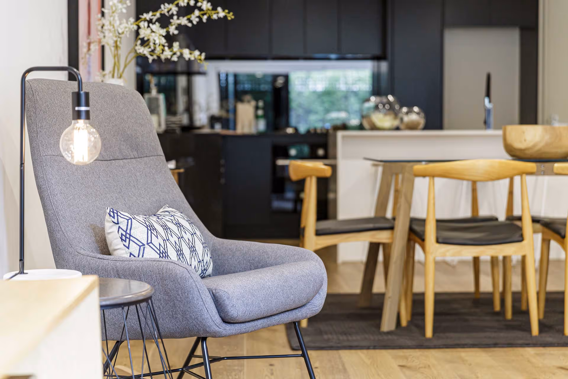 Wohnzimmer mit grauem Sessel, dekorativem Kissen, hängender Glühbirne und Holzstühlen um einen Esstisch.