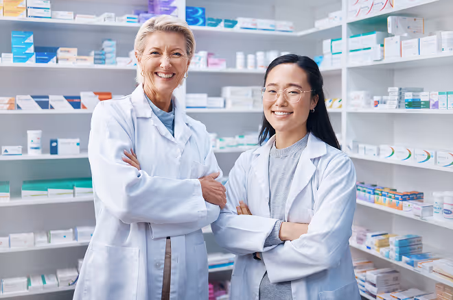 Two female pharmacists wearing white lab coats smiling with arms crossed in a pharmacy aisle.