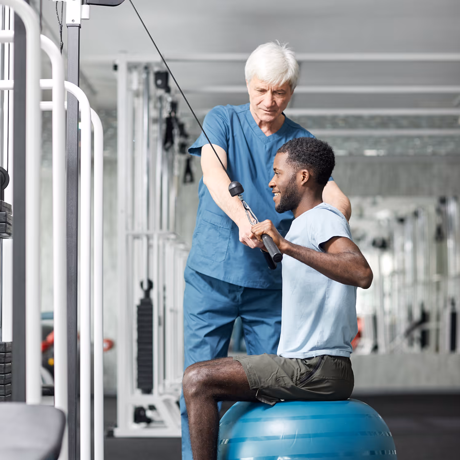 A fitness trainer assists a man seated on a blue exercise ball while using a cable machine for upper body workout.