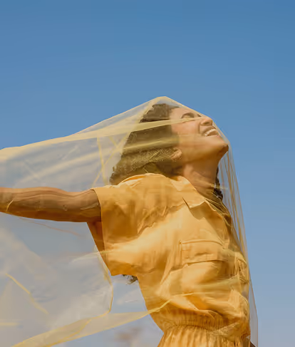 Woman in yellow dress smiling with eyes closed, arms outstretched under a sheer yellow fabric against clear blue sky.