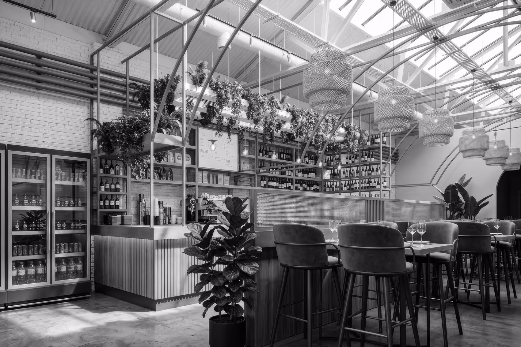 Modern café interior with high chairs, tables set with wine glasses, hanging plants, and bottles displayed behind the counter under a skylight roof.