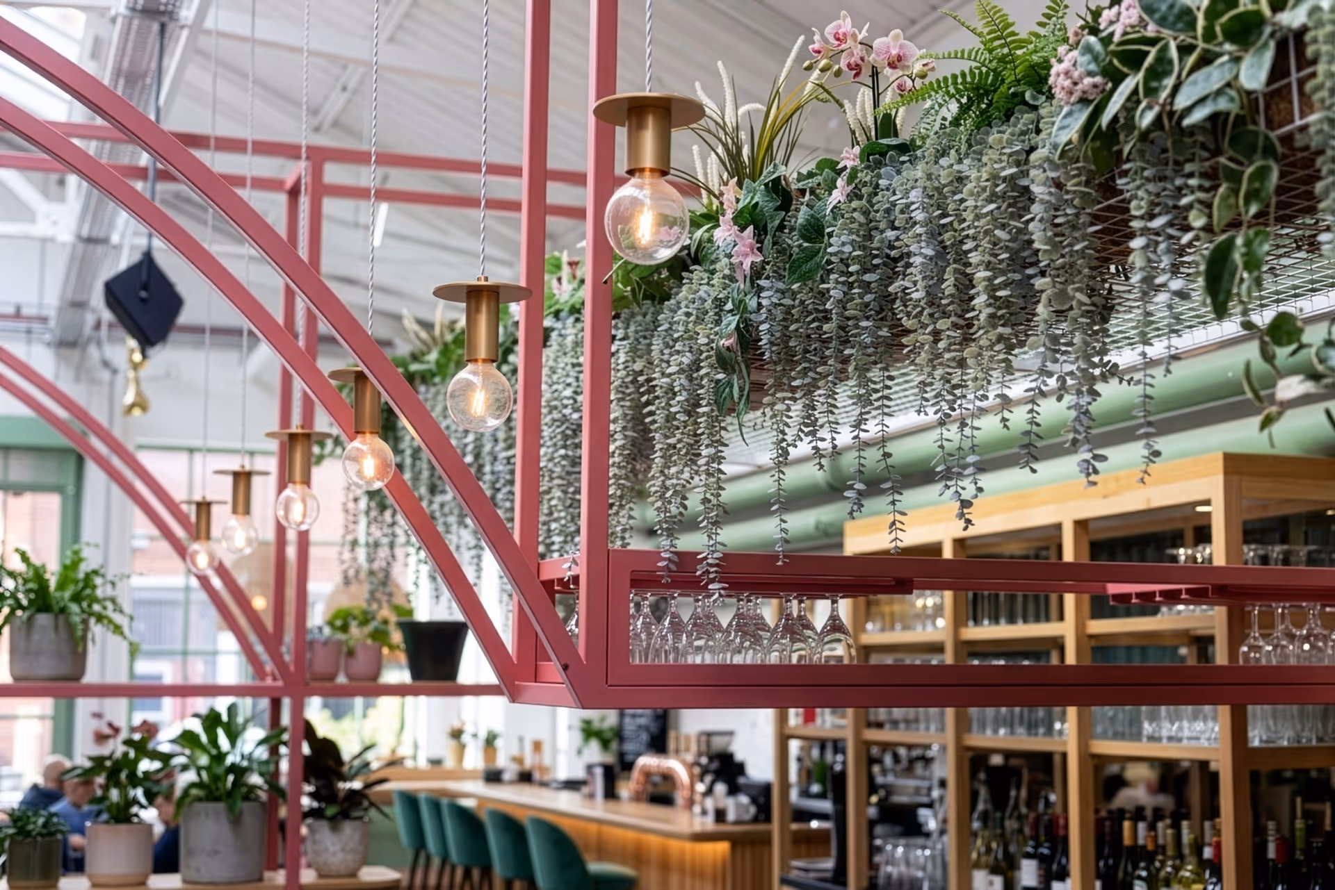 Modern bar interior with hanging plants, lit pendant lights, green bar stools, and glassware on red metal frames.