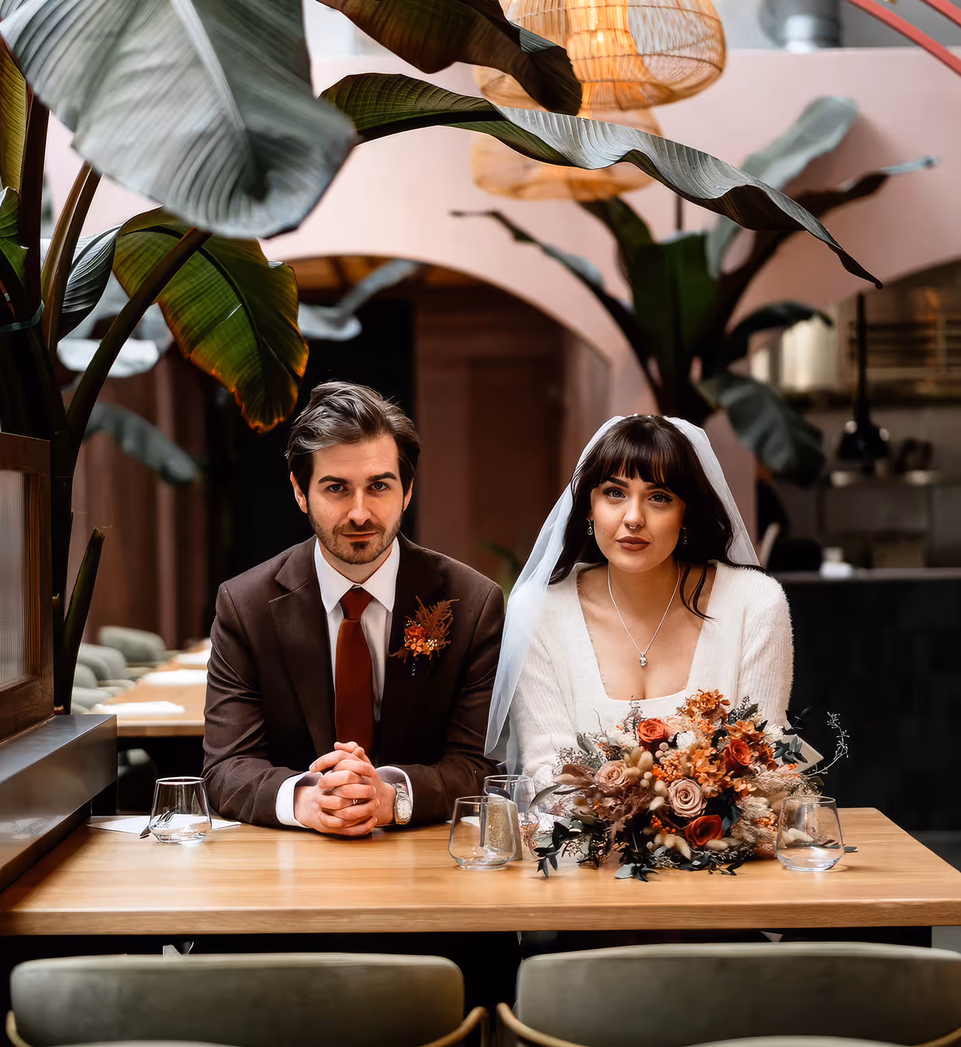 Bride in a white dress and veil with a floral bouquet sitting next to groom in brown suit and red tie at a wooden table in a modern indoor venue with large green plants.