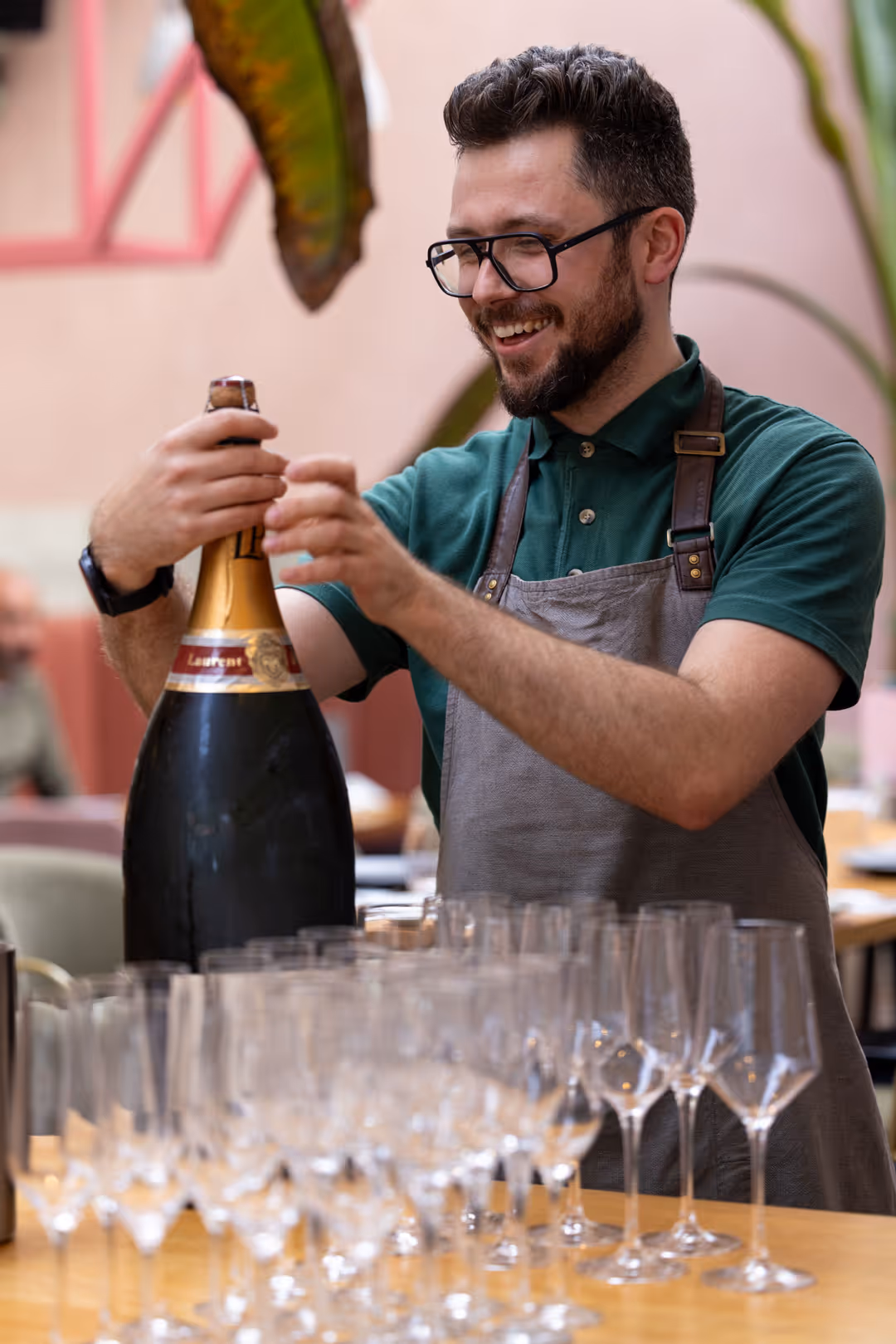 Smiling man wearing glasses and apron opening a large bottle of champagne behind a table with empty champagne glasses.