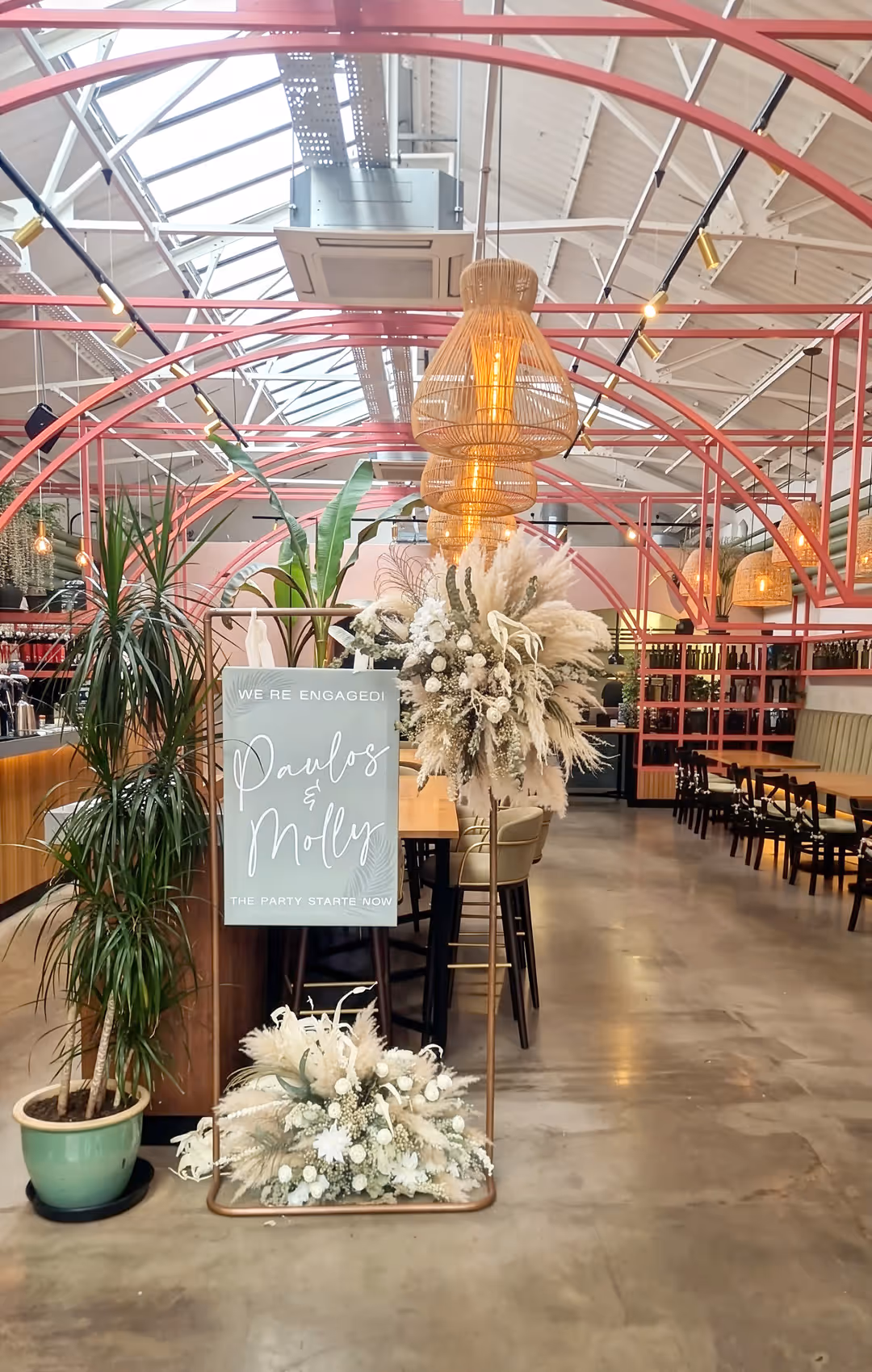 Modern event space with pink metal arches, hanging woven lamps, and a sign reading 'We're engaged! Paulos & Molly The party starts now' surrounded by floral arrangements.