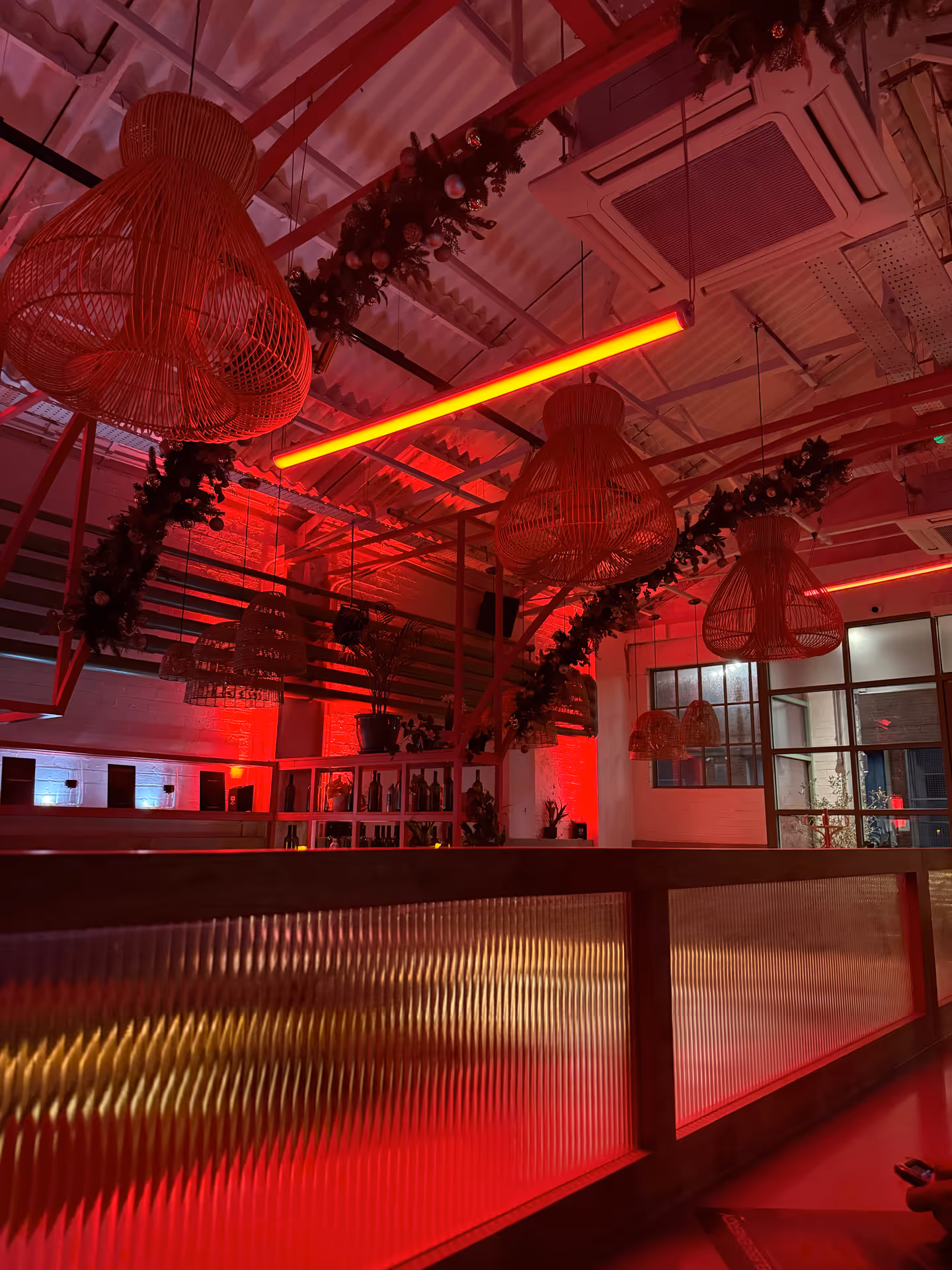 Interior of a bar or restaurant with warm red lighting, wicker pendant lamps, decorated garlands, and shelves with bottles.
