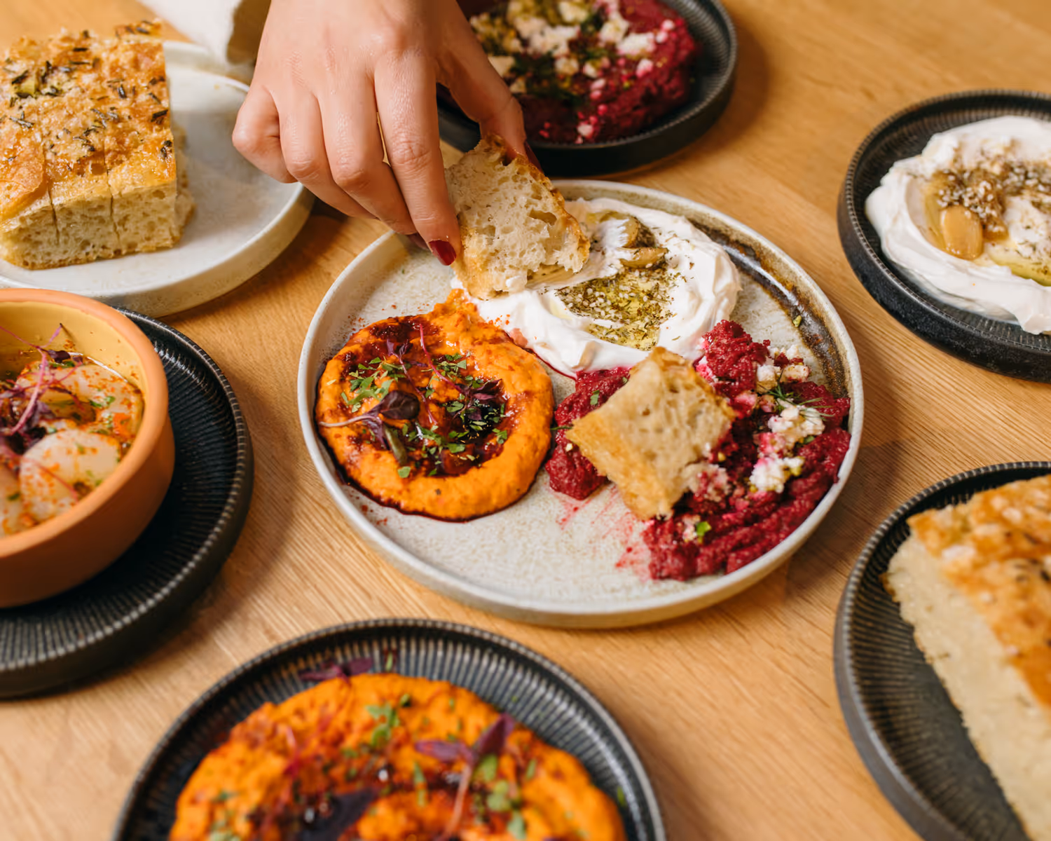 Hand dipping a piece of bread into a plate with three types of dips and additional bread and dips on surrounding plates on a wooden table.