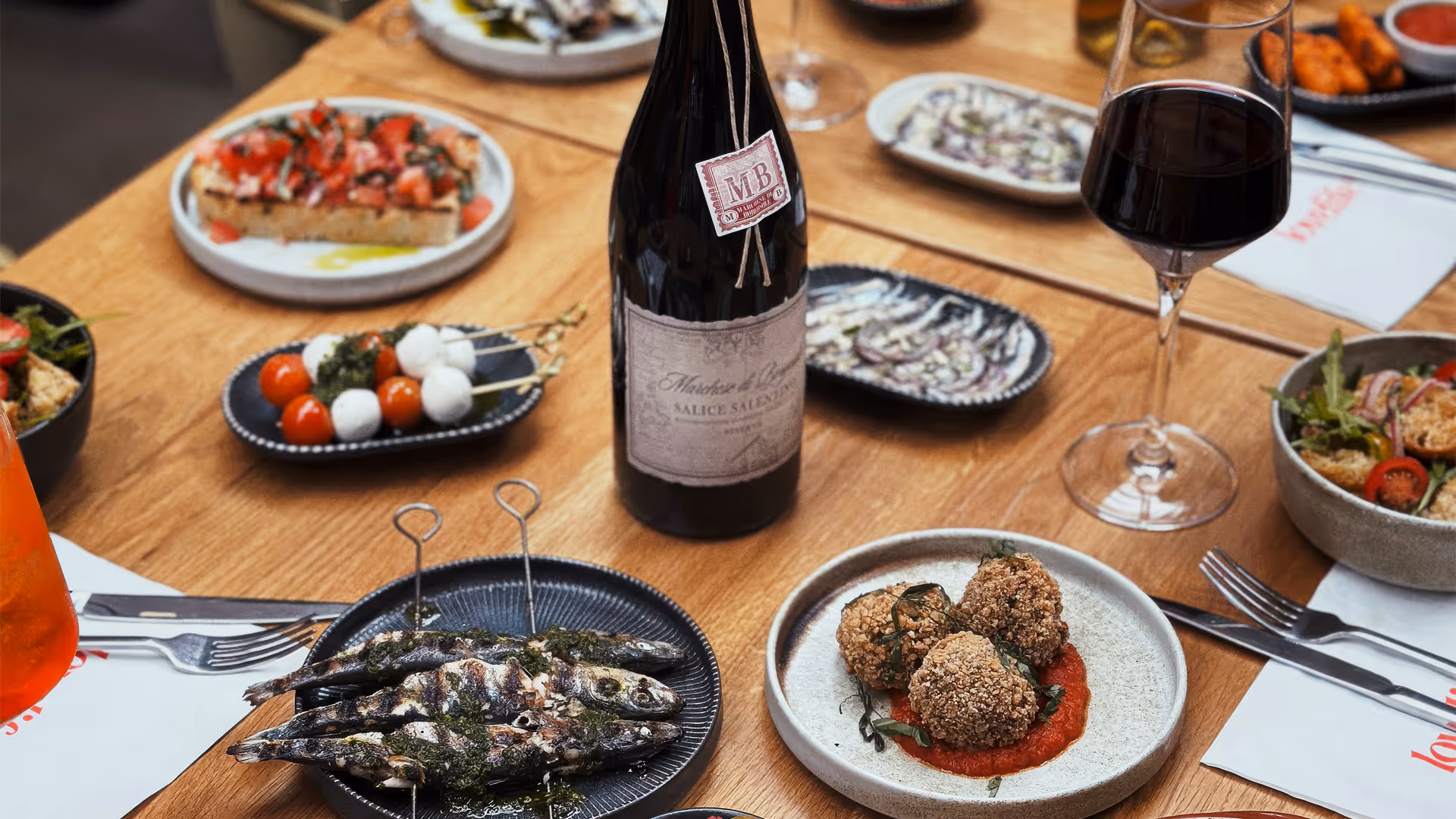 Table with various Italian dishes including grilled sardines, arancini on tomato sauce, cherry tomato and mozzarella skewers, bruschetta, salads, a bottle of red wine, a glass of red wine, and an Aperol spritz.
