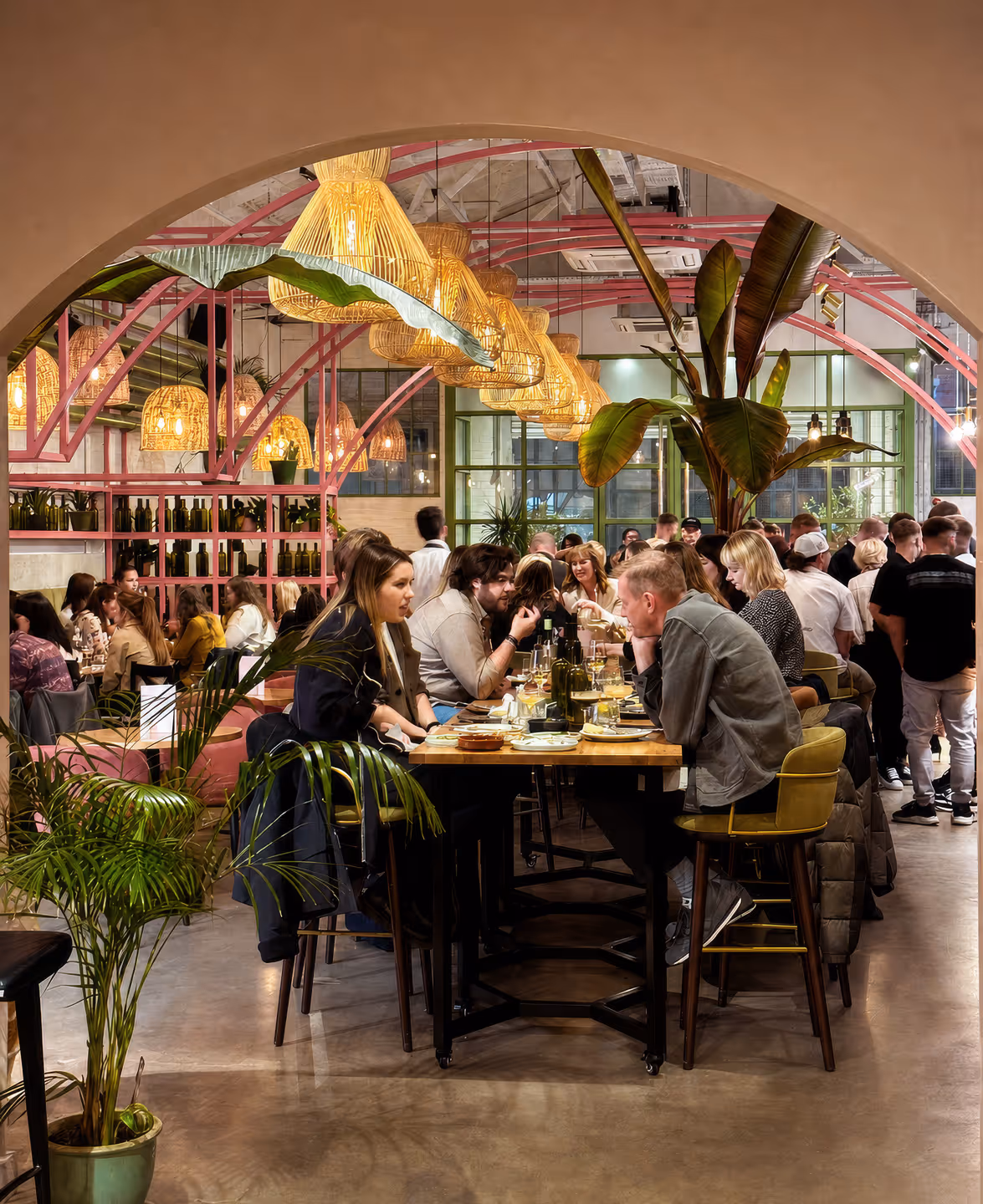 Crowded restaurant interior with groups of people dining under wicker pendant lights and pink metal arches.