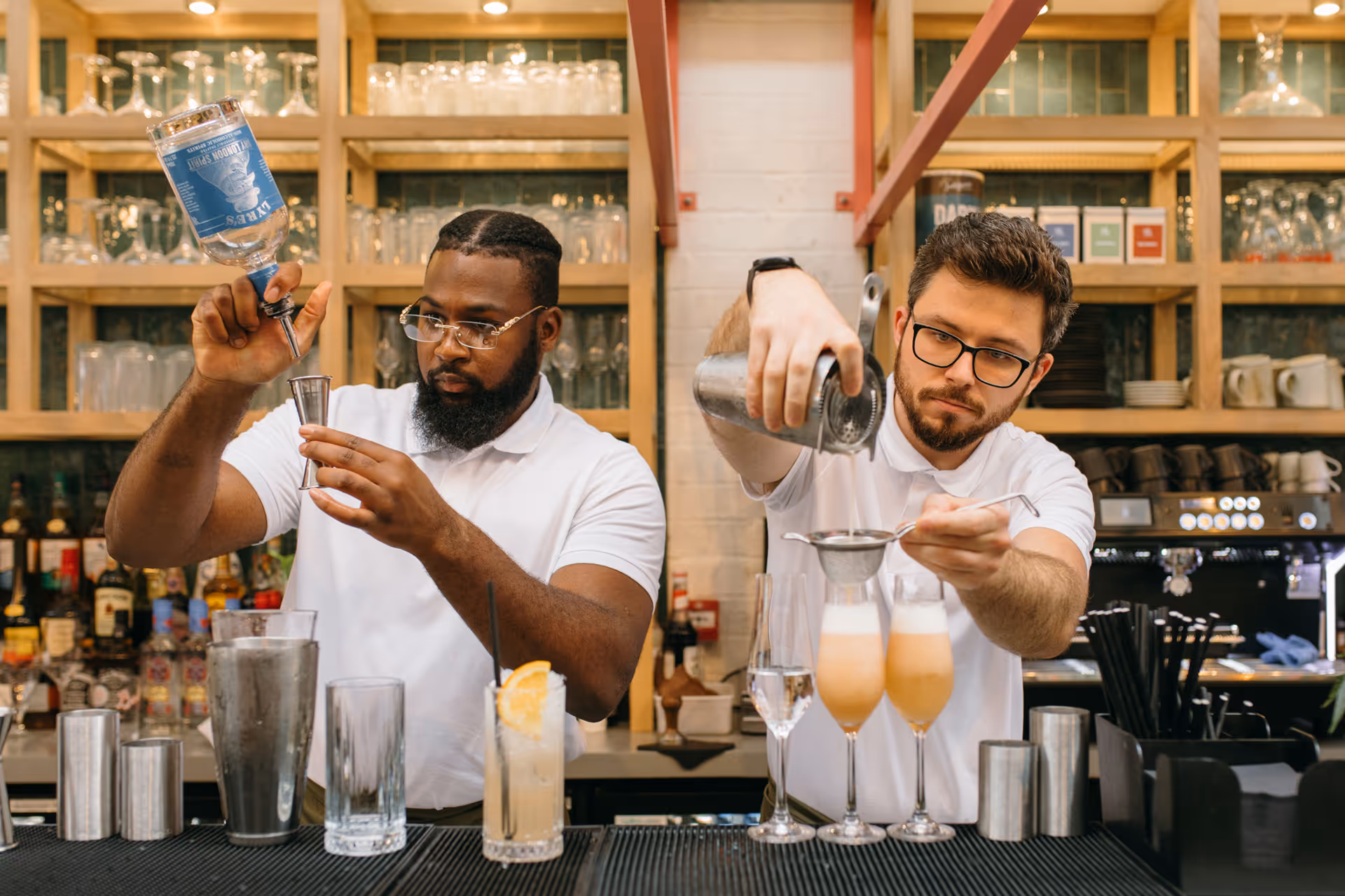 Two bartenders in white shirts preparing cocktails behind a bar with glasses and bottles on shelves.