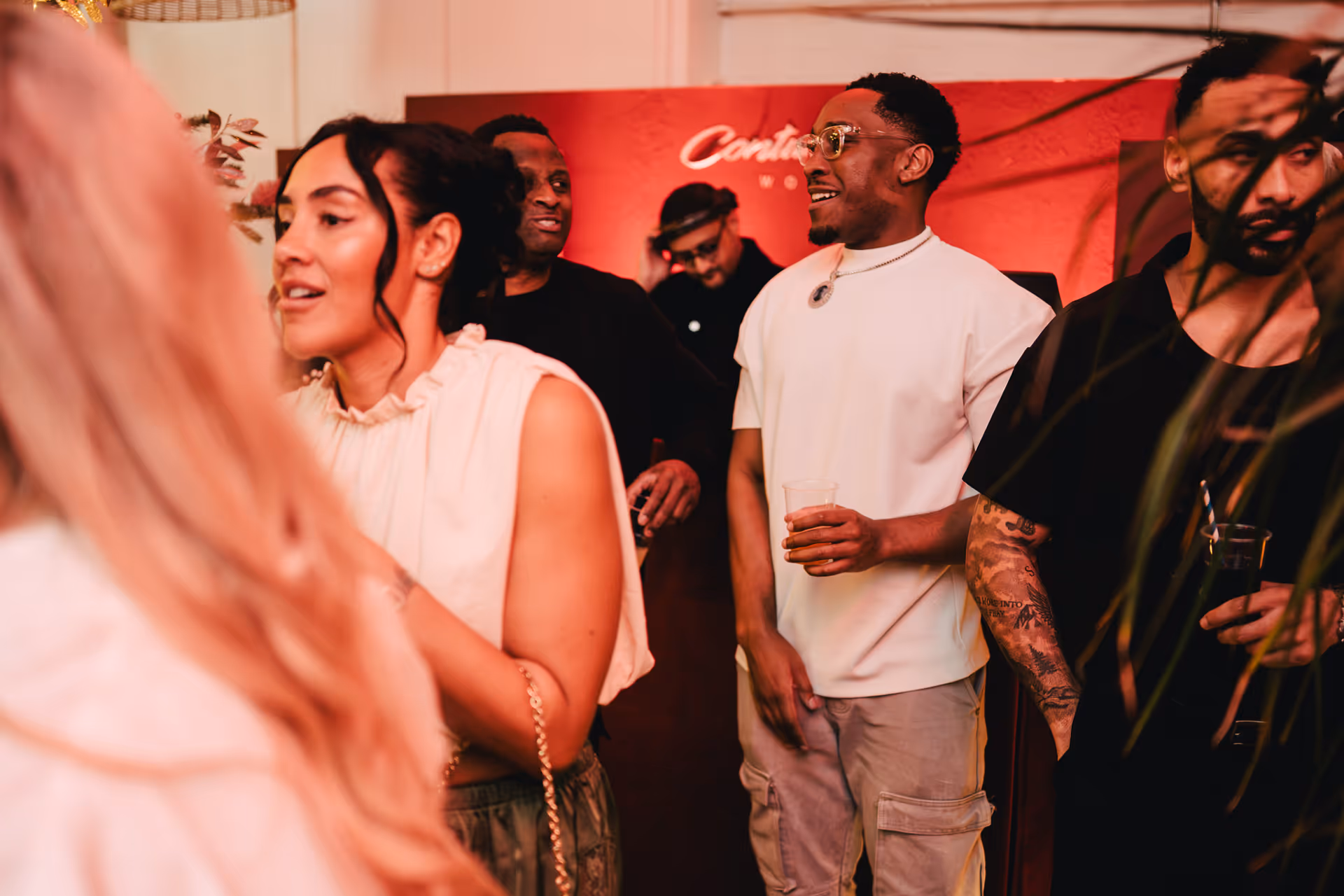 Group of people socializing indoors, some holding drinks, with a red backdrop in the background.