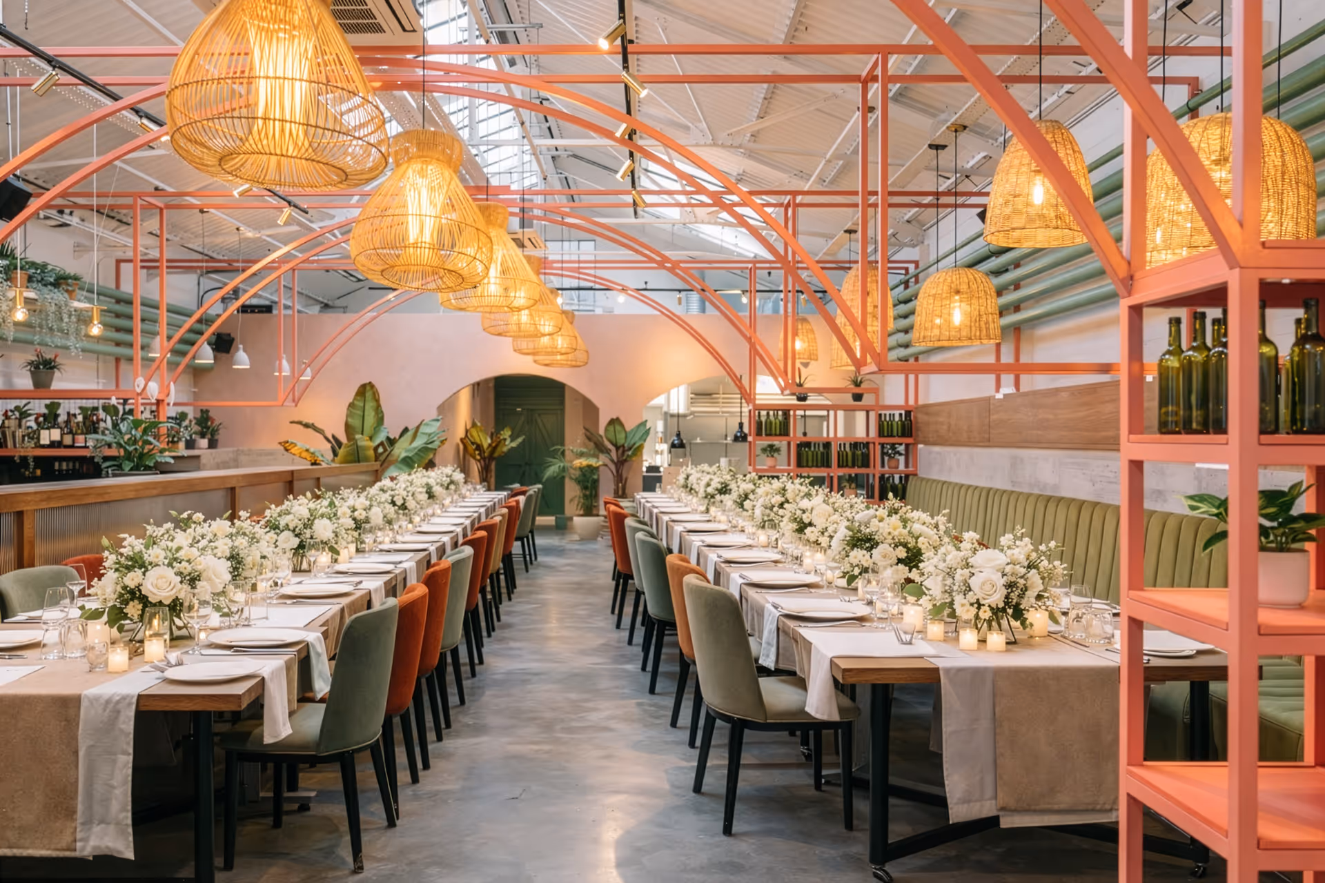 Long tables set for a wedding reception with white floral centerpieces, candles, and mix of green and rust-colored chairs under warm hanging wicker lamps.