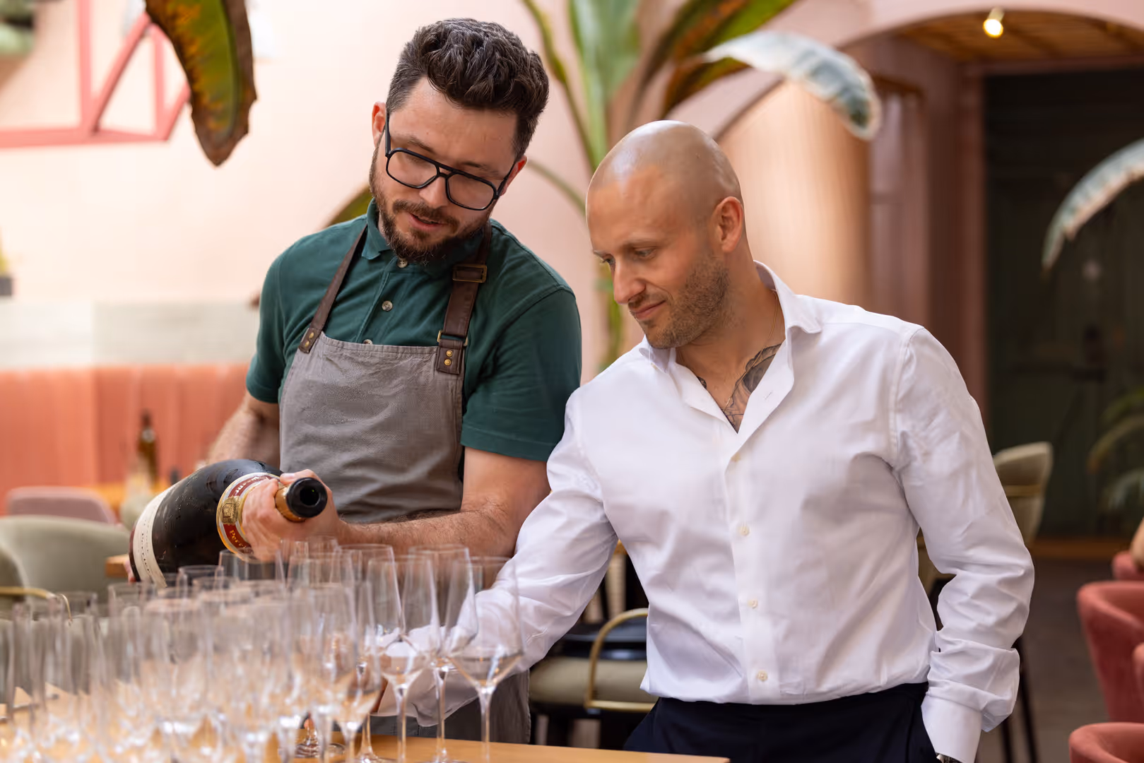 Man in an apron pouring wine into empty glasses while another man in a white shirt arranges them on a table in a restaurant setting.