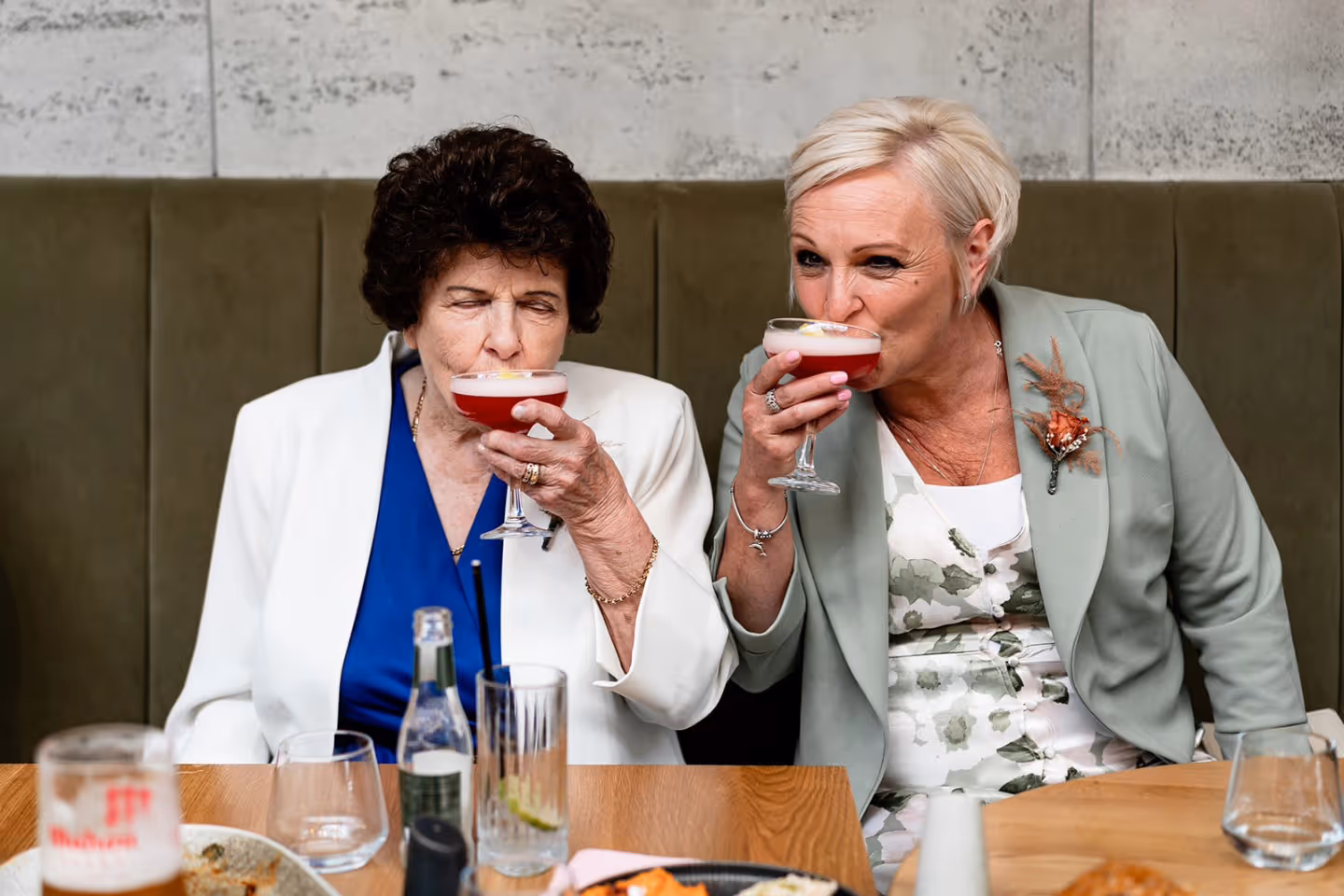 Two women sitting side by side in a booth, each drinking a red cocktail from a coupe glass.