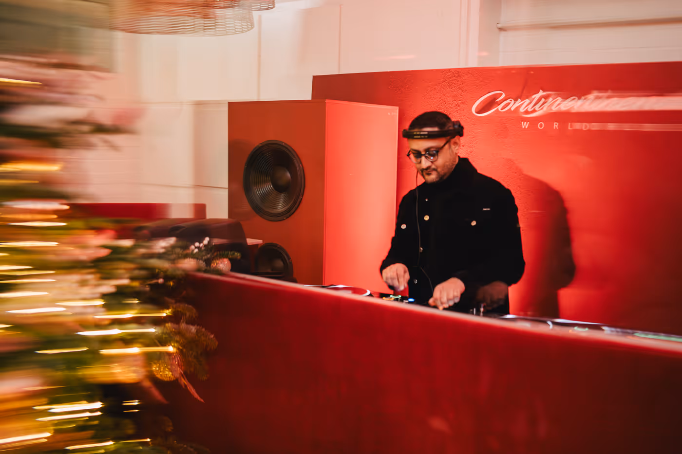 DJ wearing glasses and headphones playing music behind a red booth with a large speaker and blurred Christmas tree lights in the foreground.