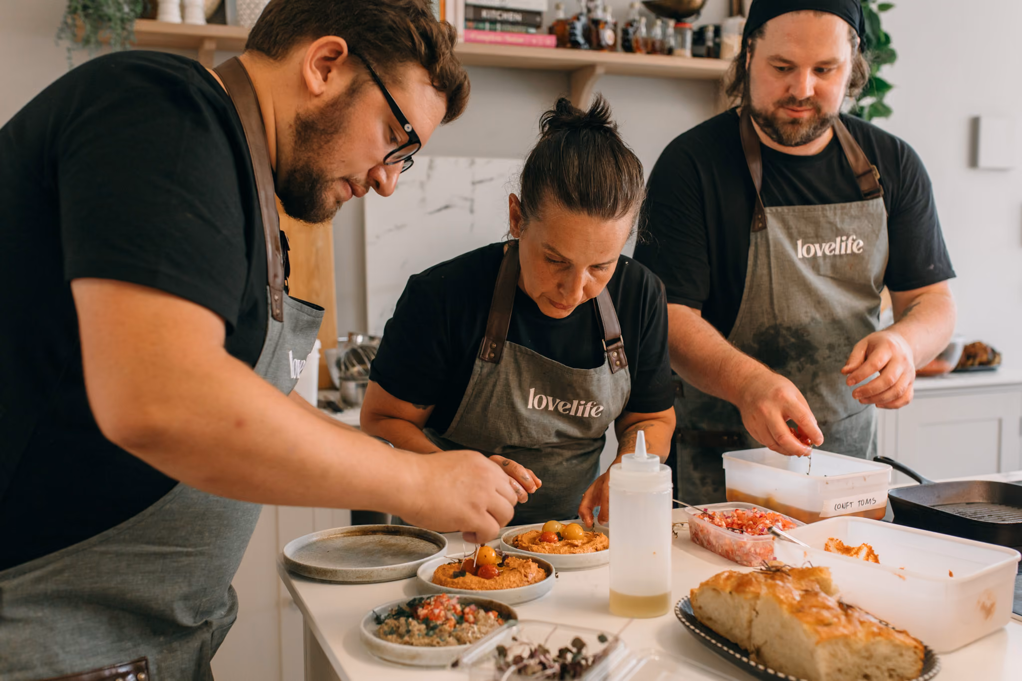 Three chefs wearing lovelife aprons preparing dishes with garnishes and ingredients in a kitchen.